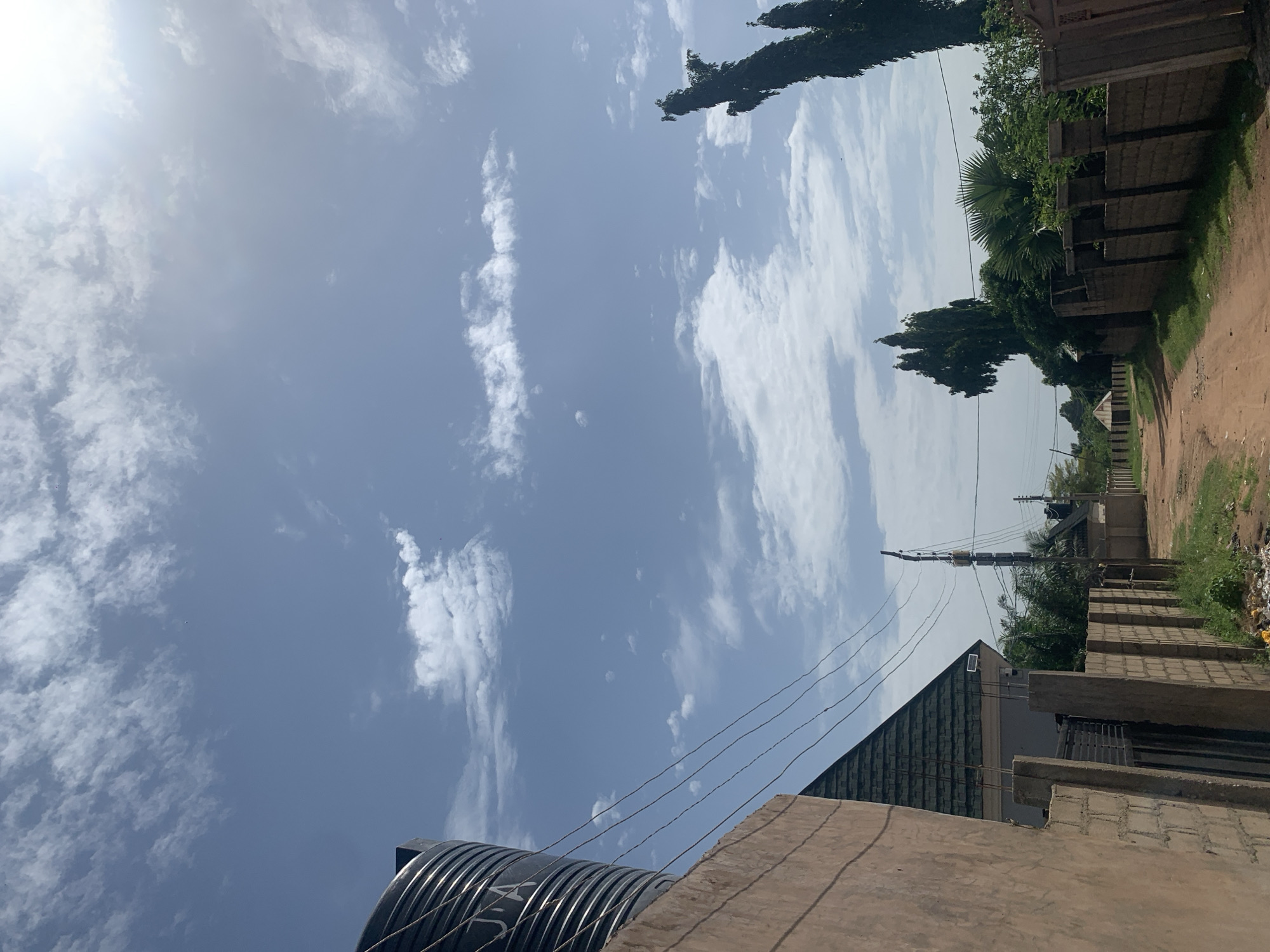 The media file depicts an outdoor scene with a prominent view of the sky filled with scattered white clouds against a clear blue backdrop. The sun is visible on the left side, casting a bright glare. In the foreground, the top of a large black water tank with metallic ribbed details is visible. There are also several thin, dark wires stretching across the frame, likely power lines or communication cables, which appear to be supported by a distant utility pole.
Below the sky and wires, the scene reveals a building with a dark, angled roof, partially obscured by another structure in the immediate foreground that appears to be under construction, with visible brickwork. The setting suggests a residential or semi-urban area in Howayi, Nigeria, as indicated by the style of buildings and the dirt road or path on the right side of the frame. Lush green trees and foliage are visible along the right edge, interspersed with a fence made of brick blocks. The overall impression is that of a bright, sunny day, possibly in the late morning or early afternoon, with a slightly hazy or overcast sky in some areas due to the scattered clouds and the intensity of the sun. No people are visible in the frame. The image is taken from a low angle, looking upwards. There is some faint white text on the water tank, but it is not clearly legible.