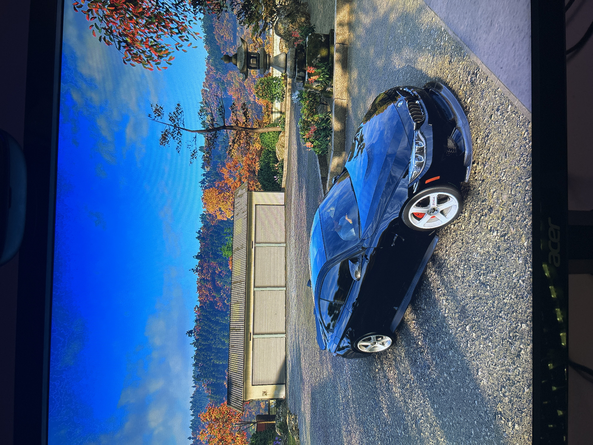 A black sports car is parked on a gravel road next to a building with white panel doors. A tree and foliage with fall colors line the left side of the road, and there is a stone lantern in the background. The sky is blue. The location is most likely a digital rendering of a scene, not Burgas, Bulgaria.