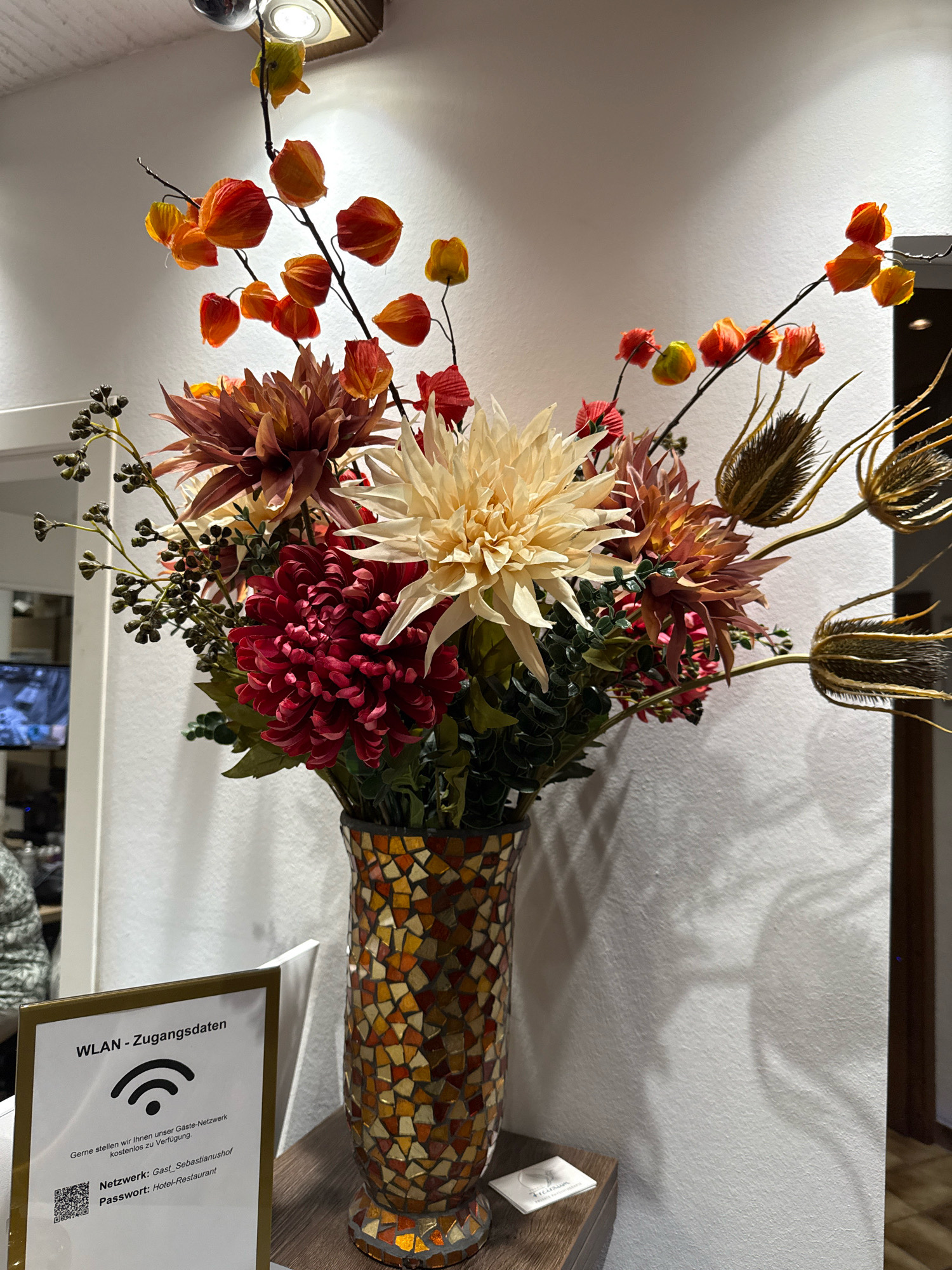 The image shows a decorative bouquet of artificial flowers in a mosaic vase placed on a wooden surface. The bouquet contains various flowers in shades of red, cream, and brown, along with dried seed pods and small berries. To the left of the vase is a white sign with the text "WLAN - Zugangsdaten" and information about a Wi-Fi network. The background is a textured white wall with shadows cast on it. The scene appears to be indoors, possibly in a hotel or restaurant lobby in Bonn, Germany, given the multilingual signage. No people are visible in the frame.