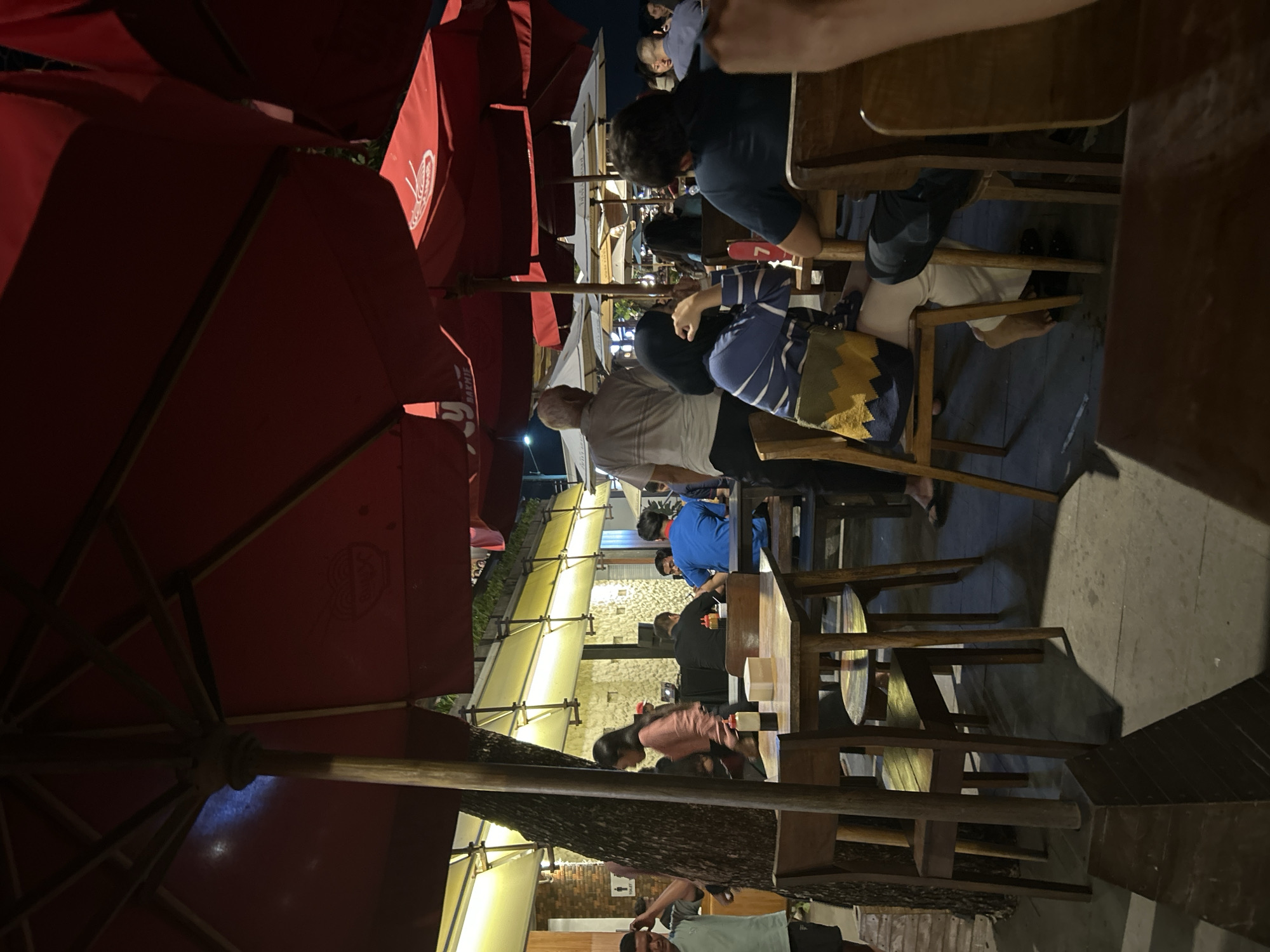 The image depicts an outdoor seating area, likely a restaurant or cafe, in Denpasar, Indonesia. Large red umbrellas provide shade over wooden tables and chairs, some of which are occupied by patrons. In the background, a building with yellow lighting and stone accents is visible, along with other outdoor seating arrangements. Several individuals are seated at tables, engaged in conversation or dining. One person, wearing a light blue shirt, is looking down at their food, while another, a woman in a blue and white striped shirt and headscarf, is looking to her left. A man in a grey shirt stands near a tree on the left side of the frame, gesturing with his hand. The overall atmosphere suggests a casual, evening dining experience.