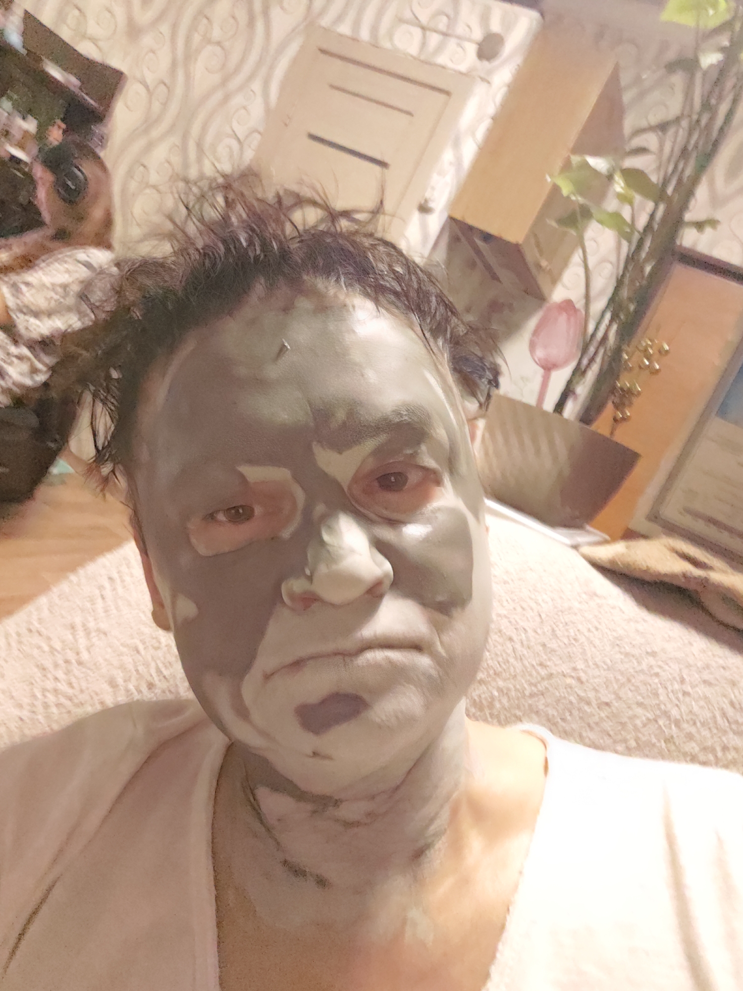 A close-up selfie shows a person with a grey mud mask applied to their face and neck. The person's hair is dark and messy, and they are wearing a light pink shirt. The background is blurred, but appears to be an indoor setting with patterned wallpaper and some shelves with objects, including a plant. There is no indication of the location being Krasnoyarsk, Russia.