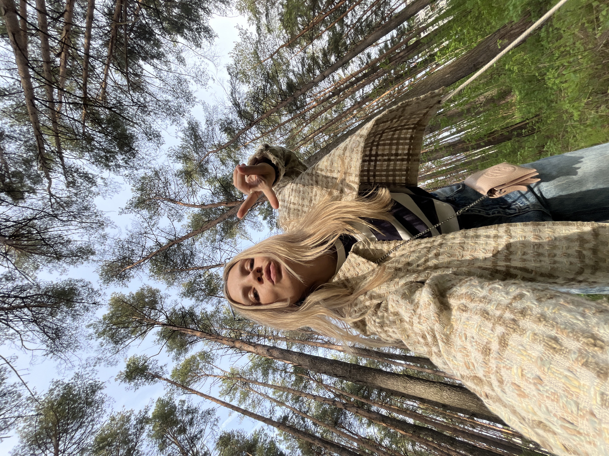 A person with blonde hair is pictured at an angle, looking at the camera while making a peace sign. They are wearing a beige jacket over a striped top and blue jeans. A small pink purse with a chain strap is visible. The background consists of tall, thin trees reaching towards the sky. The location is a forest in Chaikovsky, Russia.