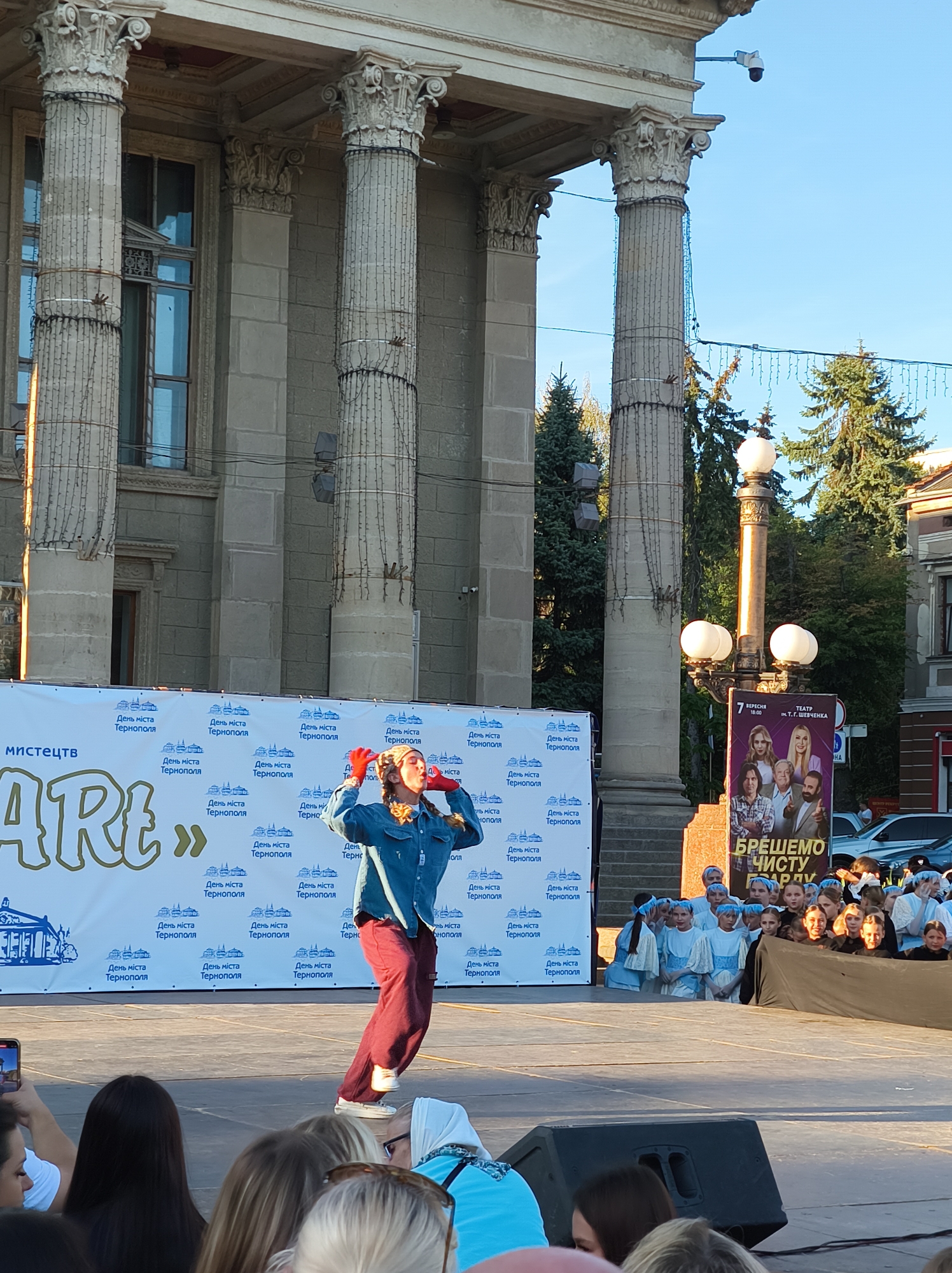 The image captures an outdoor performance event in Ternopil, Ukraine. The backdrop features a large, ornate building with classical columns, likely a public building or theater. In the foreground, a performer, dressed in a denim shirt and maroon pants, is actively engaged on a stage, with their mouth open as if singing or speaking. They are wearing red gloves. The stage is set up in front of a banner that reads "Містецтв Art" and is adorned with repeated text and logos that appear to be in Ukrainian, likely advertising the event as "День міста Тернополя" (Ternopil City Day). Behind the performer, a group of young people, some in matching light blue and white outfits, are seated and watching. To the right, a lamppost with multiple light fixtures stands prominently, and a poster advertising a performance at the T. H. Shevchenko Theater is visible. The overall scene suggests a festive community event taking place during daylight hours.