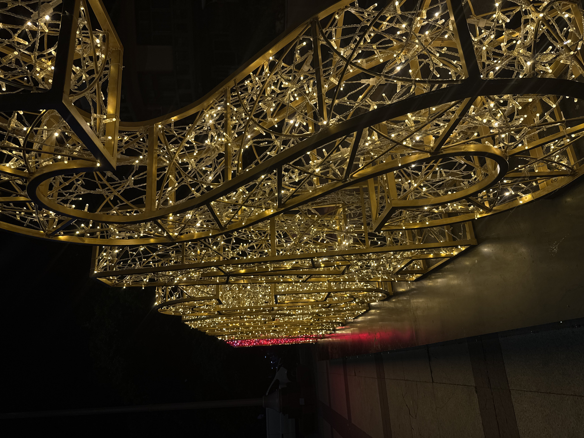 The image shows a section of a large, golden, illuminated decorative structure made of interconnected metal frames and covered in numerous small, warm white lights. The structure appears to be a large numeral or letter, possibly for a holiday or event decoration, with curved and angular sections visible. The lights create a bright, sparkling effect against the dark night sky.
To the left of the illuminated structure, a paved walkway with darker square tiles is visible, reflecting some of the light. Further to the left and in the background, a dimly lit street or plaza can be seen with a white vehicle and a lamppost. A section of the structure on the far left is illuminated with red lights.
The photograph was taken at night, likely in an outdoor urban setting. Based on the metadata associated with this image, the location is the pedestrian zone of Burgas, Bulgaria, specifically near the intersection of Bogoridi Street and Alexandrovska Street.