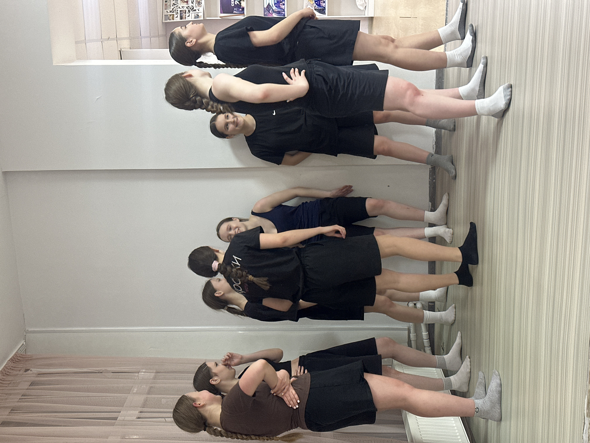 The image shows a group of seven females in what appears to be a dance or exercise studio. They are arranged in a line, standing in front of a mirror that reflects their images.
The individuals are dressed in a mix of athletic wear, including dark-colored shorts and tops. Some are wearing socks. The hair is primarily styled in braids.
The studio has a light-colored wooden floor and off-white walls. To the left, behind the women, there are semi-transparent curtains. A radiator is visible on the lower right of the image. A shelving unit is located on the upper-left part of the image, containing various objects.
While the city of Votkinsk, Russia, is mentioned, the image itself does not contain any visual elements that confirm this specific location. The scene suggests a studio environment used for activities like dance, exercise, or rehearsals.