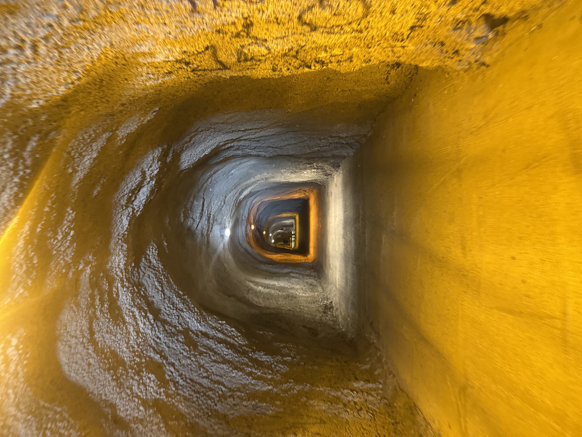 This media file captures a perspective from within a tunnel, looking towards its distant opening. The tunnel walls are rough and uneven, with textures that suggest natural rock formations or aged concrete. The lighting is artificial, casting a warm, yellowish glow that illuminates the immediate surroundings and fades into darkness further down the passage. The color palette is dominated by earthy tones of yellow and gray, creating a somewhat claustrophobic yet intriguing atmosphere.
The perspective is from ground level, looking forward into the tunnel. The immediate foreground features a textured, somewhat wet-looking surface, possibly indicating water or dampness on the tunnel floor. As the eye moves into the distance, the tunnel appears to narrow and curve, with more distinct rectangular structures and lights visible at the far end. These distant lights hint at an exit or another section of the tunnel, creating a sense of depth and mystery.
There are no visible people or specific objects clearly identifiable within the tunnel itself. The focus is solely on the architectural and textural elements of the tunnel's interior. The time of day and weather are not discernible from the image. The location context provided, Rafailovići, Montenegro, suggests this might be a natural cave, an old fortification, or a man-made passage in a coastal or mountainous region. The overall emotion conveyed is one of exploration or slight apprehension due to the enclosed and dimly lit environment. No visible text is present in the image.