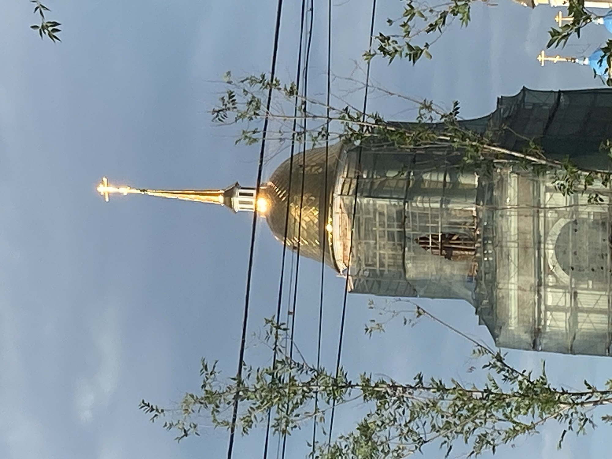 The image shows a close-up view of the dome and spire of a church, likely under renovation, as evidenced by the scaffolding and netting covering a portion of the structure. The dome is golden and highly reflective, catching the light. Atop the spire is a bright, glowing cross. Several trees with green foliage are visible in the foreground, partially obscuring the view of the church. Power lines are also strung across the image. In the background, the sky is overcast with a mix of gray and lighter blue tones. The scene suggests an urban environment, possibly Petropavl City Administration, Kazakhstan, given the context provided.