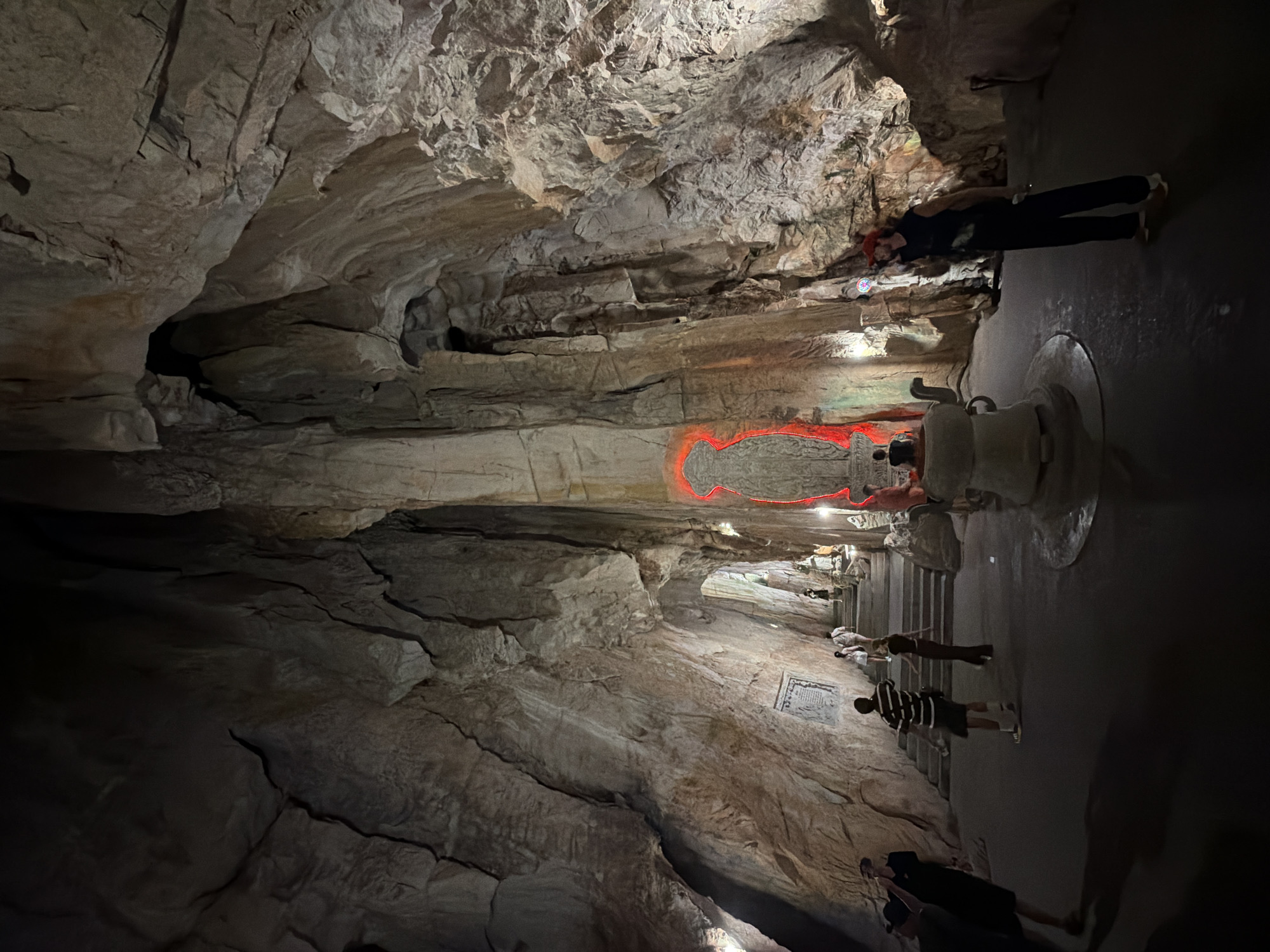 A dimly lit cave with textured rock formations is shown. A group of people is inside the cave, walking on a path. Some people are standing near an illuminated area, possibly observing a display. The cave walls are rugged and uneven, with visible layers and crevices. There is a red outline of a map, or other marking, on the cave wall. It is Son Tra Cave, Da Nang, Vietnam.
