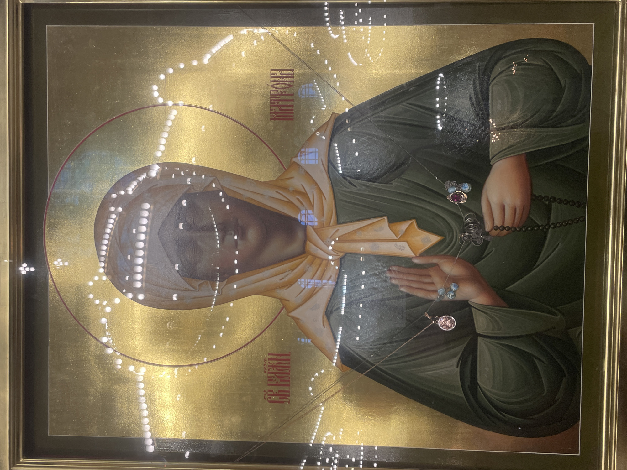 This is a framed icon depicting a woman with a gold background and a halo. The woman is wearing a light yellow headscarf and a dark green robe. She has her hands clasped in front of her, holding a rosary. There are red inscriptions on either side of her head. The icon is likely a religious artifact, possibly housed in a church or museum. The image is highly reflective, indicating it is behind glass, and the reflections show lights and possibly architectural details. Without further context, the specific location in Kronstadt, Russia, cannot be determined from the image alone.