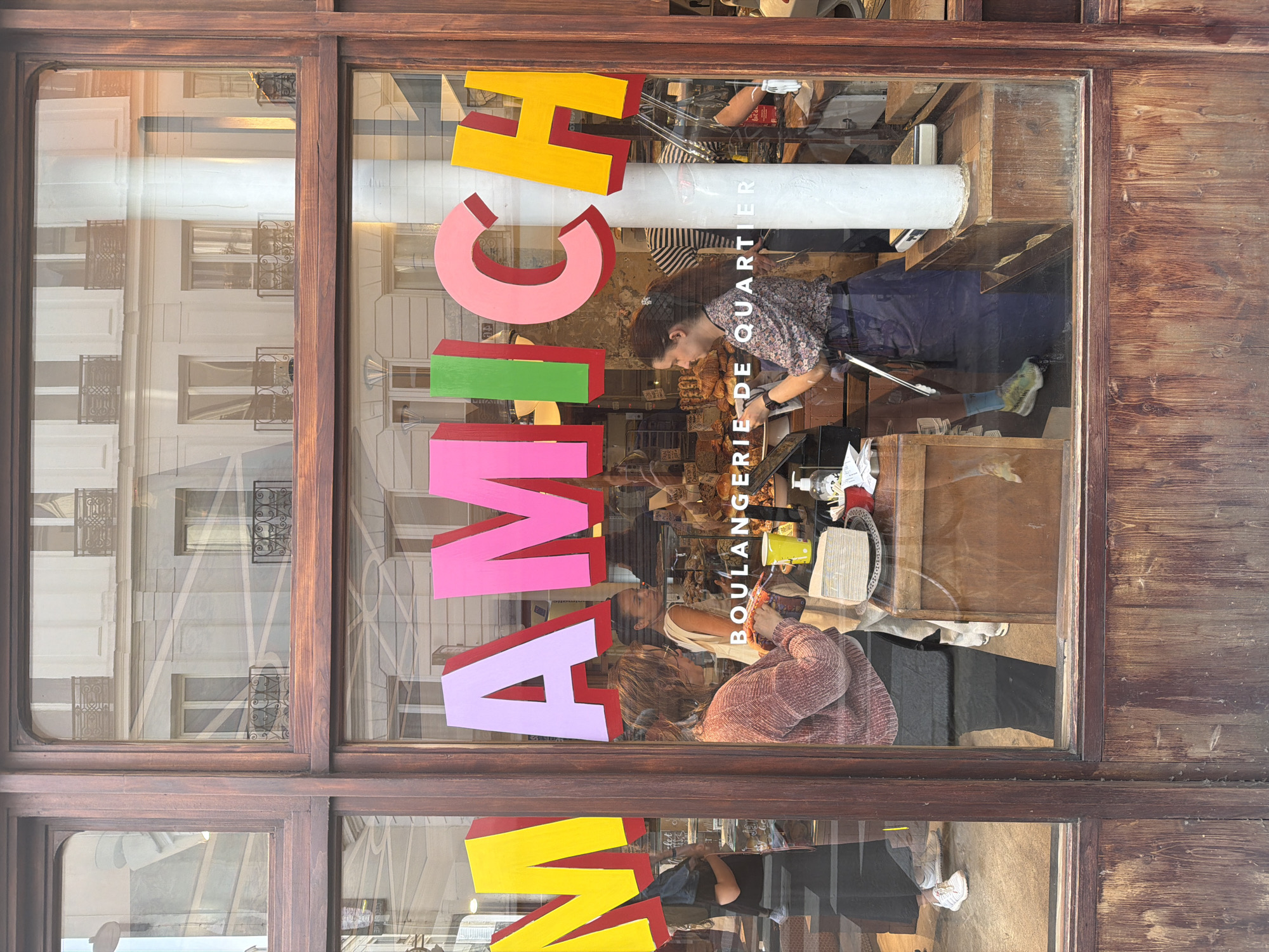 A street-level storefront in Paris, France is viewed through its window. The window is divided into sections by a dark, wood frame. Multi-colored 3D block letters spelling "AMICH" are affixed to the window, in three sections, top, middle and bottom. The letters are in yellow and red, pink, green, and lavender. Inside the store, at least three people are visible. One person in a dark, floral-patterned shirt and blue apron is working at a counter. Behind that person, in the upper right quadrant, another person is visible in a striped shirt. A third person in a salmon-colored sweater is visible in the lower portion of the window. Loaves of bread are visible on the counter. The words "BOULANGERIE DE QUARTIER" are printed vertically on the glass near the middle right side of the window. The buildings across the street are reflected in the glass. The wood on the storefront window frame appears worn.