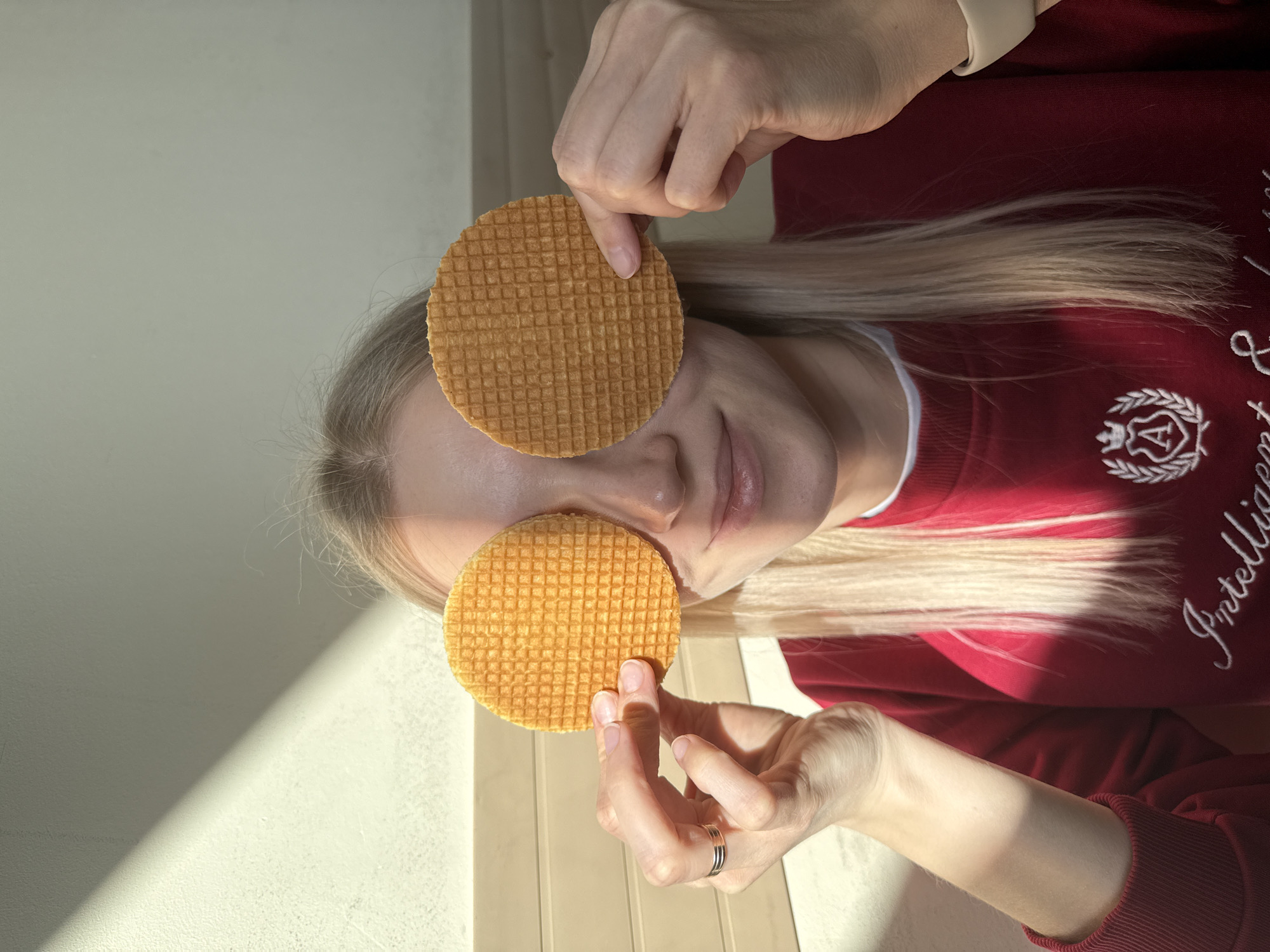 A person with long blonde hair is holding two stroopwafels over their eyes in an indoor setting. The person is wearing a red sweatshirt with white writing on the right sleeve and a white collar. The background consists of a white wall and a tan surface. The light source is coming from the left, creating a shadow on the wall and surface. There is no explicit indication of the location in Izhevsk, Russia.