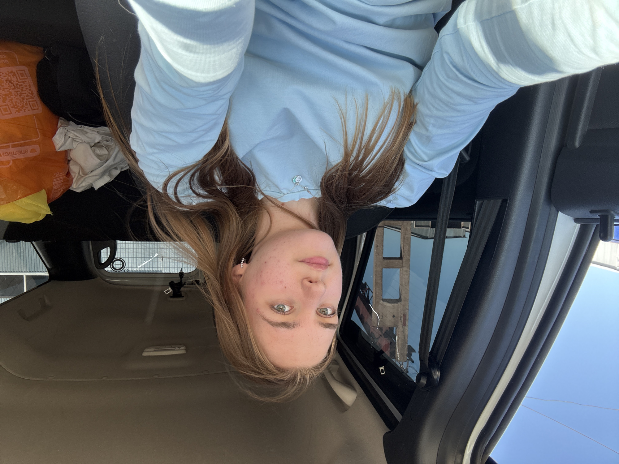 The image shows a young woman with long brown hair and light blue eyes, wearing a light blue long-sleeved shirt, inside a car. The woman is upside down in the frame, suggesting she has taken the photo herself from an unusual angle. In the background, visible through the car window, there is a building in Perm, Russia. An orange grocery bag and some white clothes are in the back.