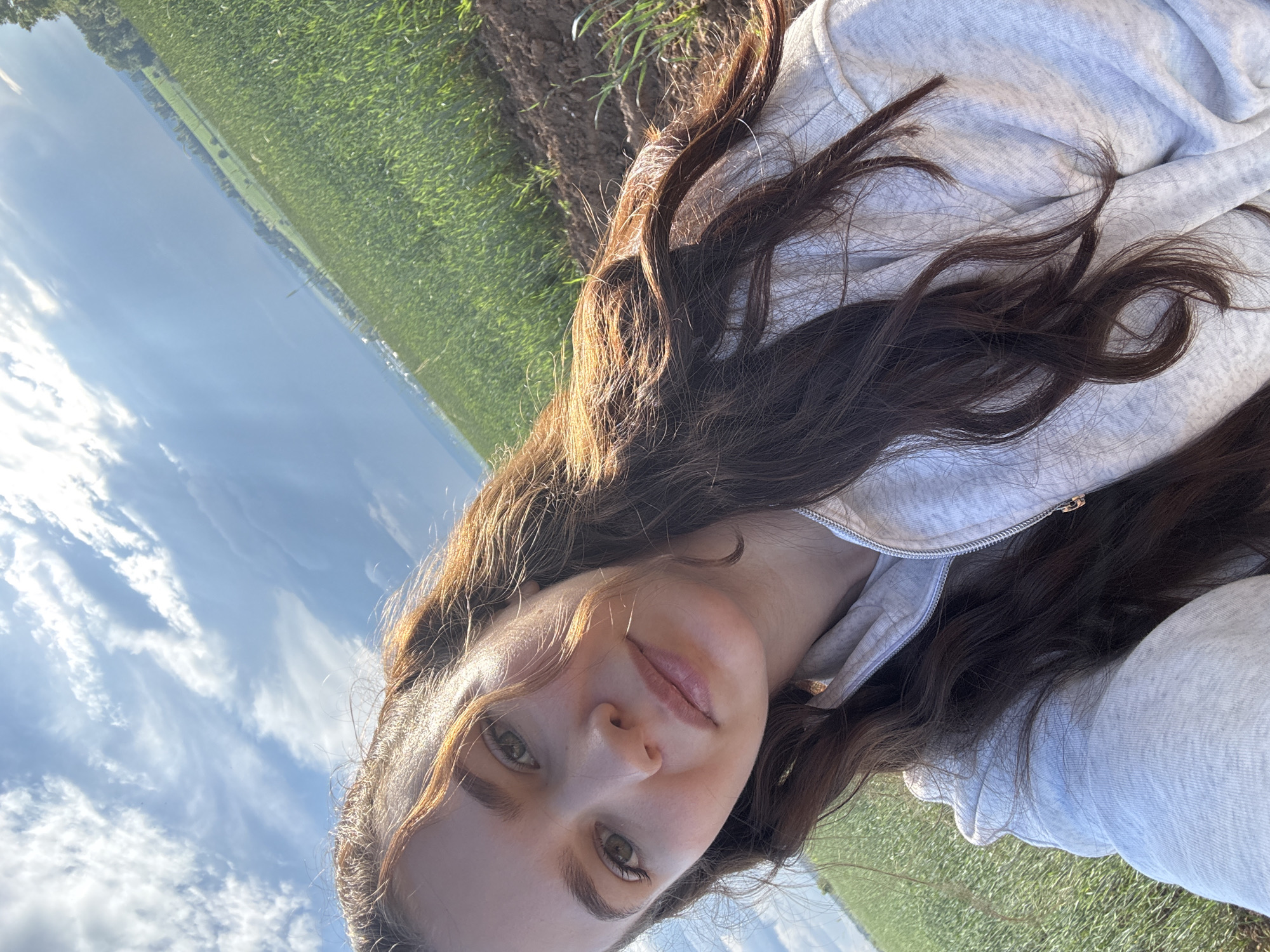 A young woman with long, dark brown hair and light brown eyes is captured in a selfie outdoors. She is wearing a light gray zippered hoodie. Her hair is partially pulled back, and strands frame her face. The background shows a bright blue sky with white clouds and a lush green field, suggesting a rural or agricultural setting. The lighting indicates it is daytime, likely sunny. The image is cropped to focus on the woman's upper body and face, with the landscape serving as a backdrop. There is no specific information available about the location being Lukintsy, Russia.