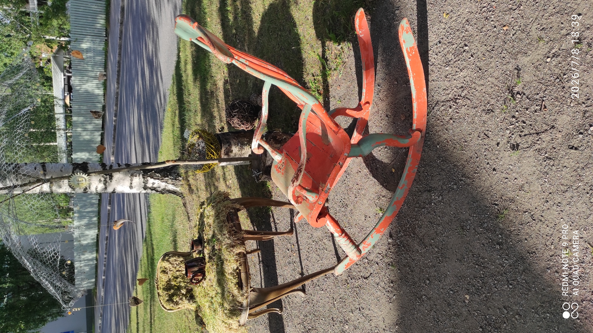This image depicts an outdoor scene in the city of Iisalmi, Finland. In the foreground, a weathered, orange and teal rocking chair sits on a gravel surface. The chair has a distressed paint finish, showing signs of age and wear. To the left of the rocking chair is a small, ornate table covered in moss, with a brown ceramic mug and cup placed on top. Behind the table, a birch tree stands prominently, adorned with a decorative wire mesh canopy and several hanging ornaments, including heart shapes and a sun face. The background shows a residential street with a light-colored wooden fence and trees. The overall setting suggests a whimsical, outdoor art installation or decorative display.