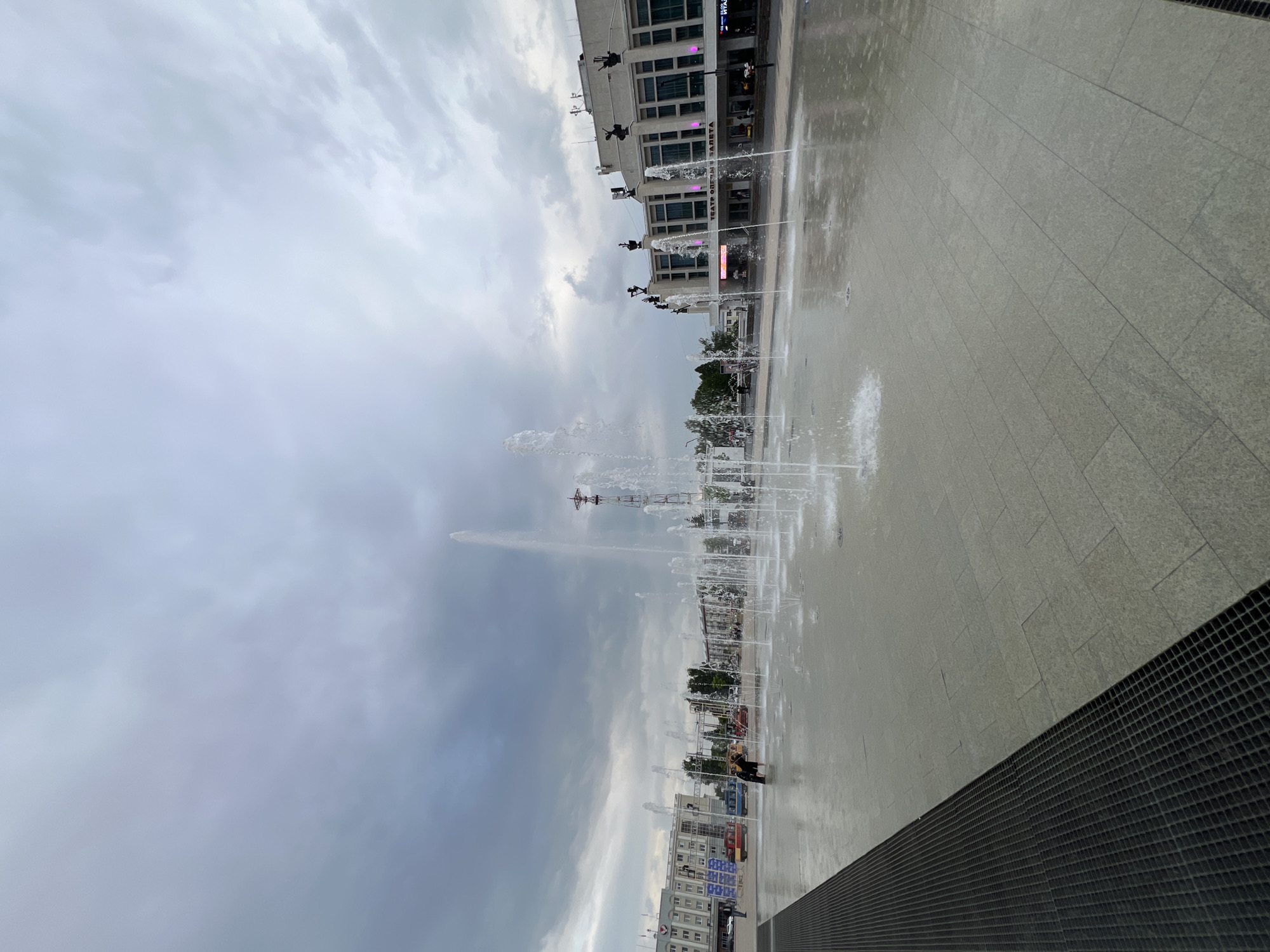 The image depicts a public square with a fountain, possibly located in Izhevsk, Russia. The scene is captured on an overcast day, suggested by the muted light and cloudy sky. The ground is paved with large, light-colored tiles, and a section of the area is wet, indicating recent or ongoing activity from the fountain.
Several jets of water from the fountain are visible, spraying upwards into the air. In the background, a modern building with multiple windows and architectural embellishments stands under the cloudy sky. Some figures can be seen near the building, though they are small in the frame. A prominent sign on the building displays text, "ТЕАТР ОПЕРЫ И БАЛЕТА," which translates to "Opera and Ballet Theater." There are also some smaller structures and trees visible further in the distance.
Towards the bottom right of the image, a dark, gridded structure runs diagonally across the frame, potentially a drainage grate or a decorative element of the square's design. The overall impression is of a somewhat grey and cool atmosphere, consistent with an overcast day. The presence of the fountain suggests it is either a warm season or the fountain is operated regardless of the temperature for aesthetic purposes. No specific time of day can be determined due to the uniform lighting.