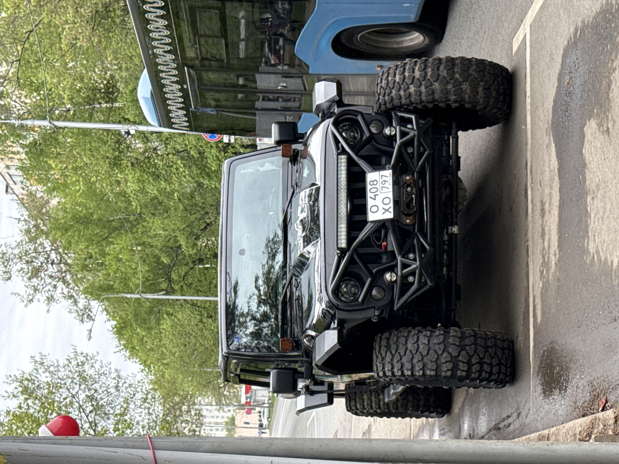 The image shows a black Jeep Wrangler parked on a street in Moscow, Russia. The vehicle has a custom front bumper and a large light bar. The license plate number is O 408 XO 797 RUS. A blue bus is partially visible behind the Jeep. Trees and buildings are visible in the background. The street surface appears wet, with a visible white painted line. A red and white bollard is in the foreground.