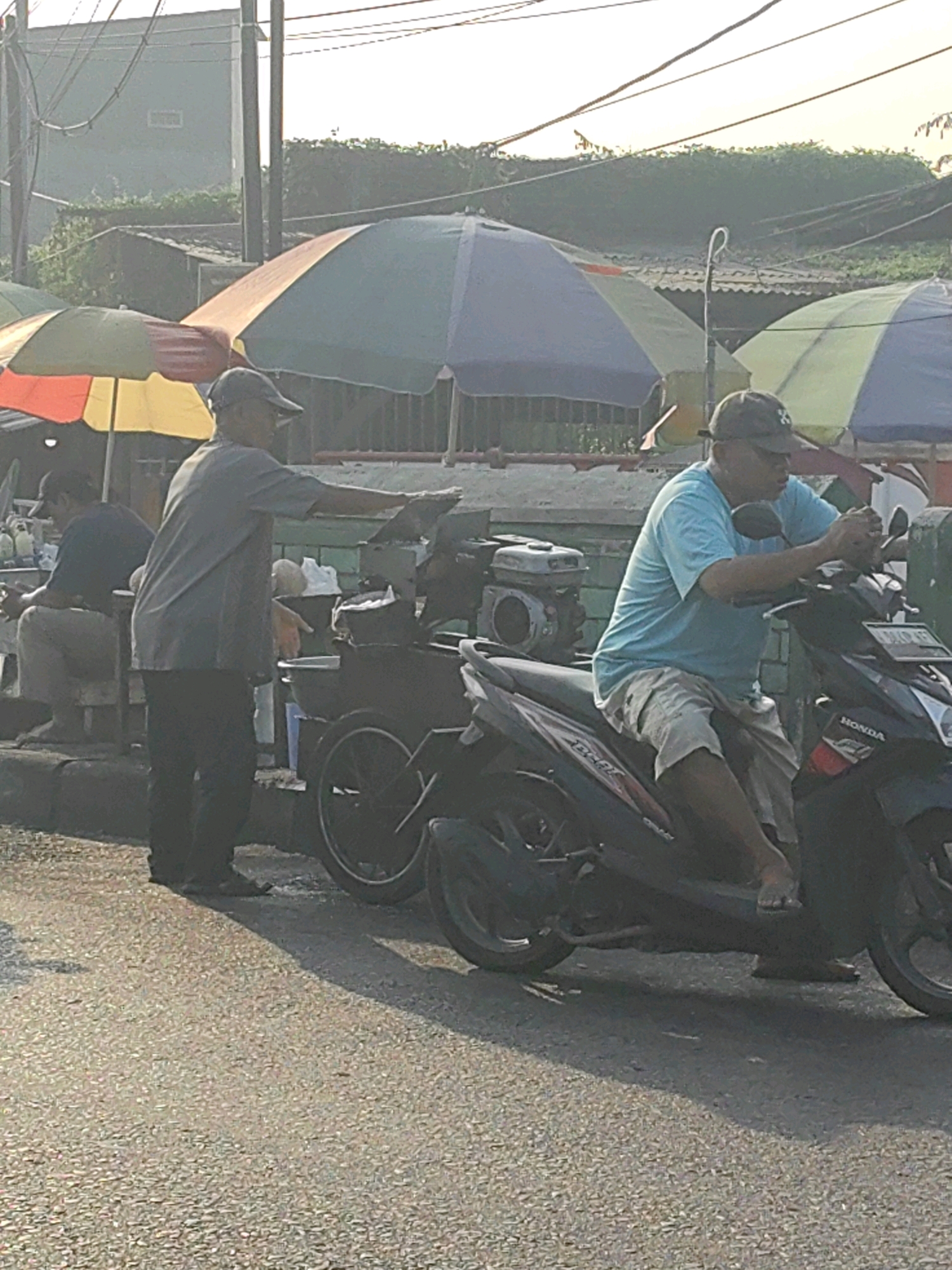 In the city of Keboansikep, Indonesia, a scene unfolds with two men and a street food stall. A man, wearing a gray t-shirt and dark pants, stands behind a portable food cart, extending his left arm and hand towards the cart, as if attending to a customer or preparing food. He wears a dark baseball cap. To his right, another man is seated on a dark-colored motorcycle. He is wearing a light blue t-shirt, light-colored shorts, and a baseball cap. He holds the handlebars of the motorcycle with both hands. The motorcycle itself is parked on the side of a paved road, near the food stall. Behind the food stall, several colorful umbrellas, likely for shade, are visible, along with a building and some vegetation. The lighting suggests it is daytime, possibly morning or late afternoon, with soft sunlight casting shadows. A third person is partially visible sitting on a stool at the far left, also facing the street food stall.