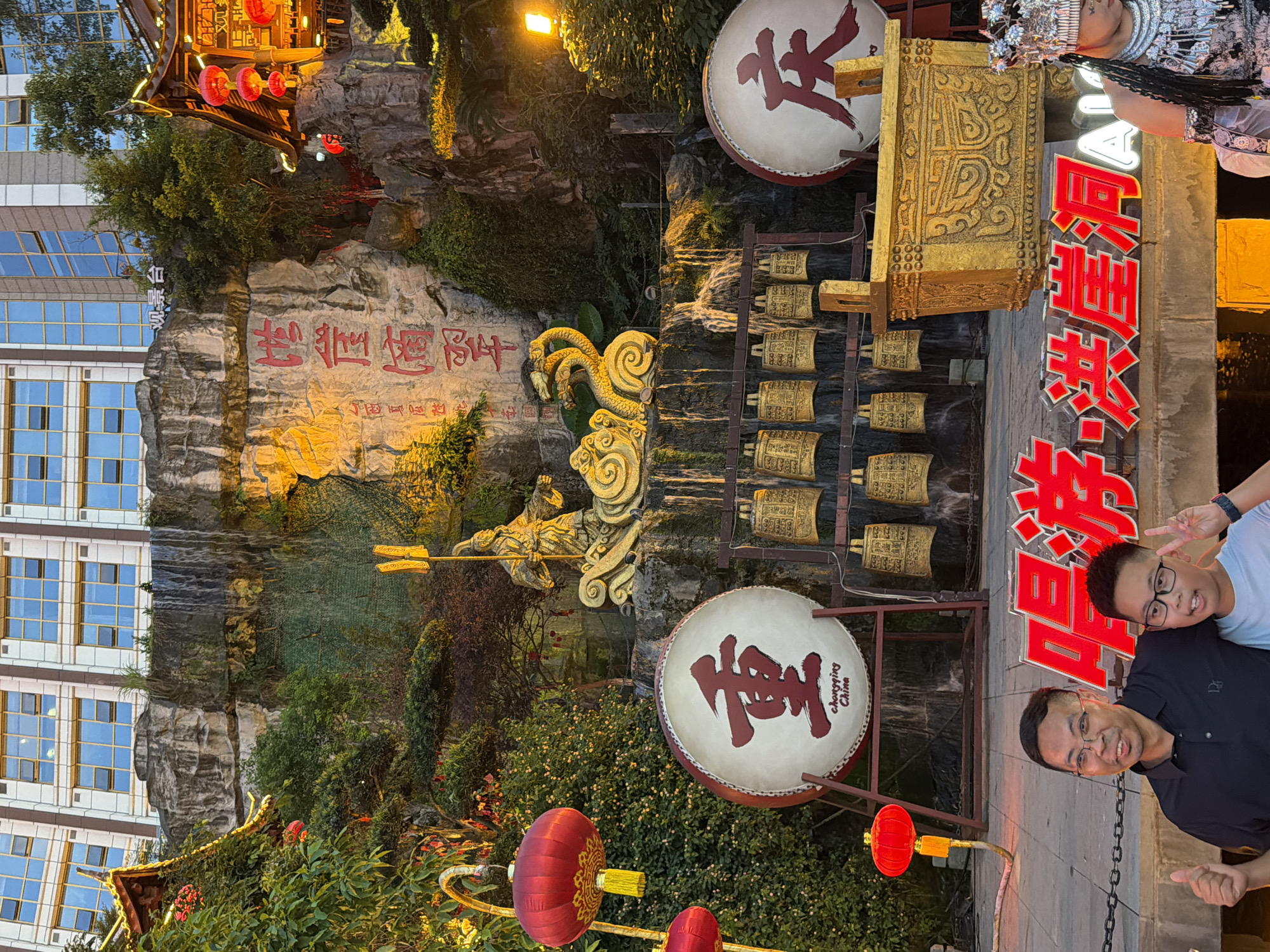 In Yuzhong District, China, a scene unfolds with traditional Chinese cultural elements. A large, illuminated waterfall cascades down a rocky cliff face adorned with red Chinese characters. At the base of the waterfall, a collection of golden bells hangs, and a golden ornate chest sits nearby. Two large drums, one with the Chinese character "重" and the other with "庆", are prominently displayed. The drum on the left also features "Chongqing China". Two men pose for a photo in front of a red neon sign. The man on the left, wearing glasses and a black polo shirt, gives a thumbs-up. The younger man on the right, also wearing glasses, holds up a peace sign. In the background, a modern building with many windows stands tall, contrasting with the traditional structures and decorations. Red lanterns hang from branches, adding to the festive atmosphere. A person in ornate traditional attire, with a silver headdress and jewelry, is partially visible on the right side of the frame.