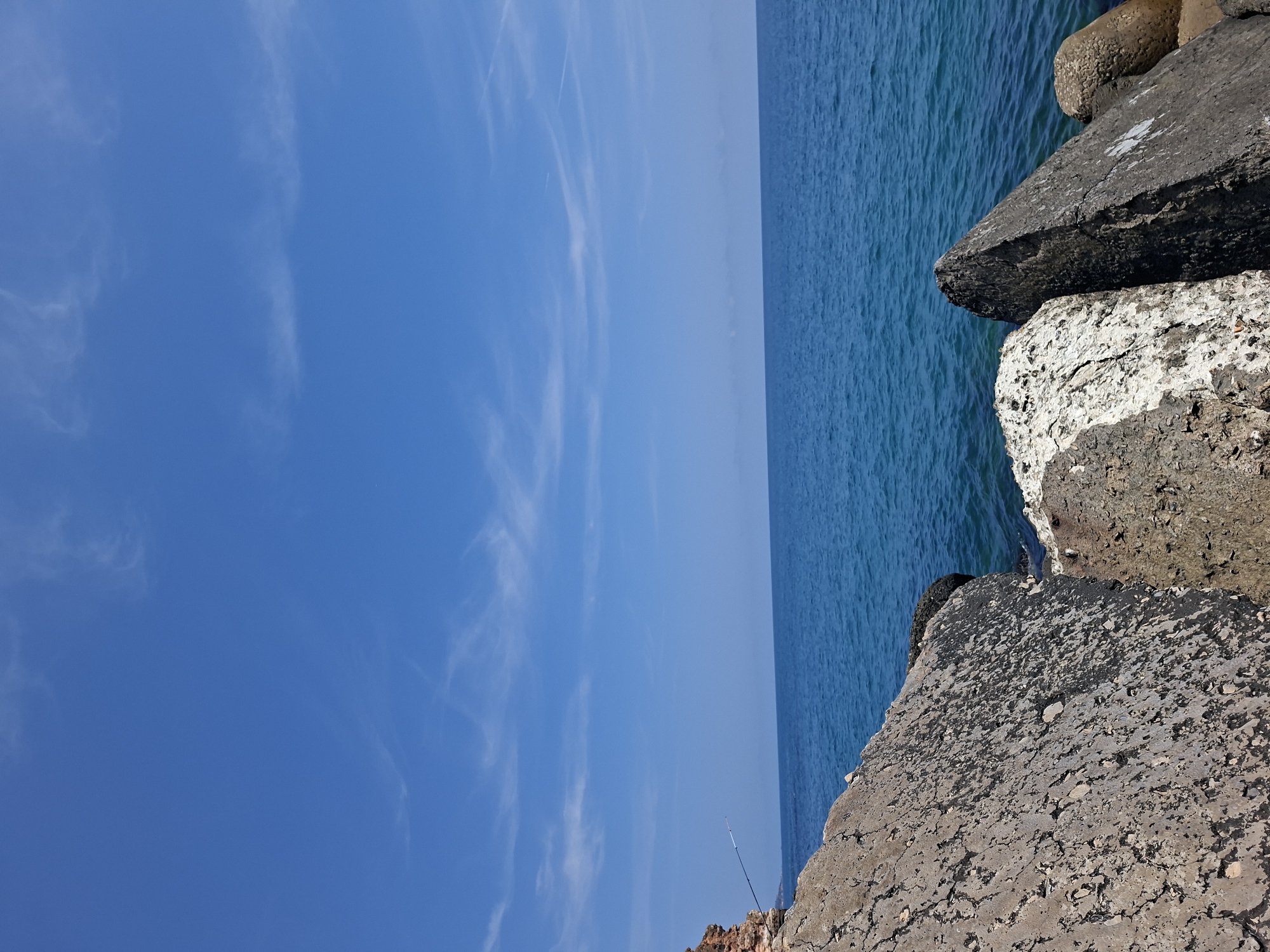 A still, medium-shot, eye-level photograph captures a coastal scene in Balgarevo, Bulgaria, under a bright, clear blue sky dotted with wispy white clouds. The vast expanse of the deep blue sea dominates the horizon, with gentle waves creating a textured surface. In the foreground, large, weathered rocks and concrete breakwaters are piled along the shoreline, their rough textures and varying shades of grey and brown contrasting with the smooth sea and sky. A thin fishing rod extends from a rocky outcrop on the left, suggesting a solitary angler engaged in the activity. The overall atmosphere is one of tranquil solitude and natural beauty.