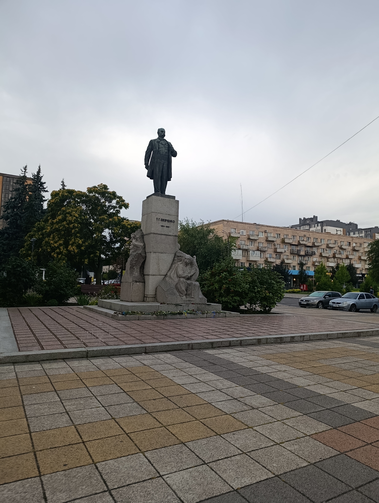 The image depicts a statue of Taras Shevchenko, a prominent Ukrainian poet, located in the city of Cherkasy, Ukraine. The statue is situated on a granite pedestal inscribed with "Т.Г. Шевченко 1814-1861". The statue itself is a full-body bronze sculpture of Shevchenko standing in a formal suit. The pedestal is adorned with bas-relief sculptures depicting figures associated with his work. The statue is located in a paved square with a patterned walkway in the foreground. In the background, there are trees, a multi-story apartment building with balconies, and cars parked on a street. The sky is overcast.