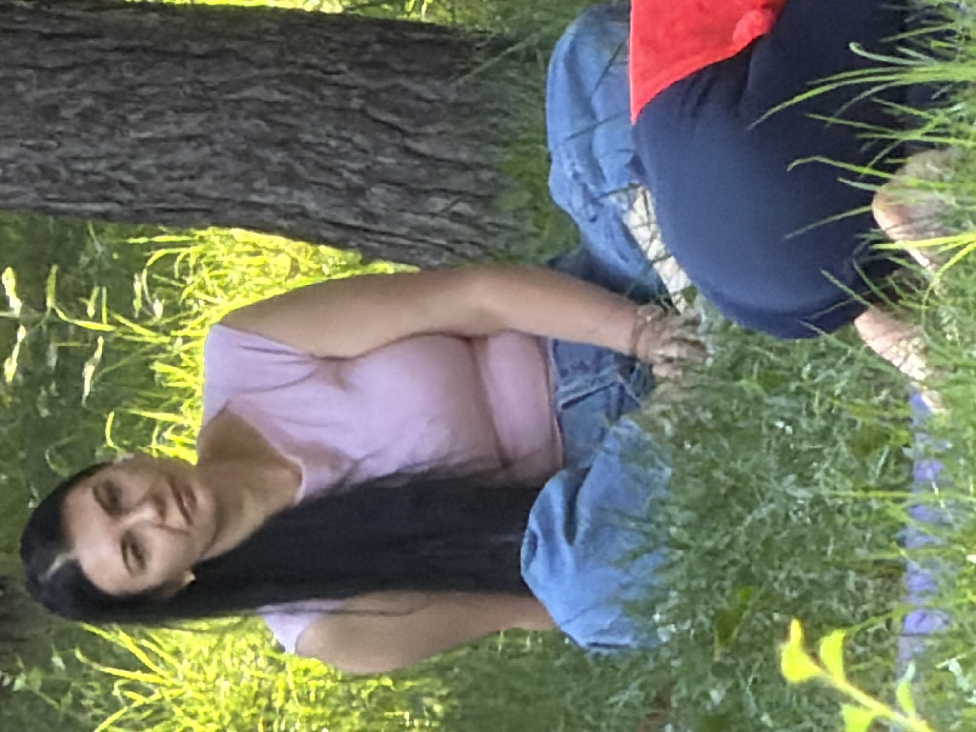 A woman with long black hair and fair skin, wearing a light pink t-shirt and blue jeans, sits on the grass in a park. Her hair is pulled back in a ponytail. She is looking to her right, with her arms resting on her lap. Behind her, a large tree trunk is visible. In the foreground, another person is kneeling on the grass, wearing a red shirt and dark blue pants. The ground is covered in green grass and some fallen leaves. The scene appears to be a casual outdoor gathering in a park setting, likely during daytime given the bright lighting.