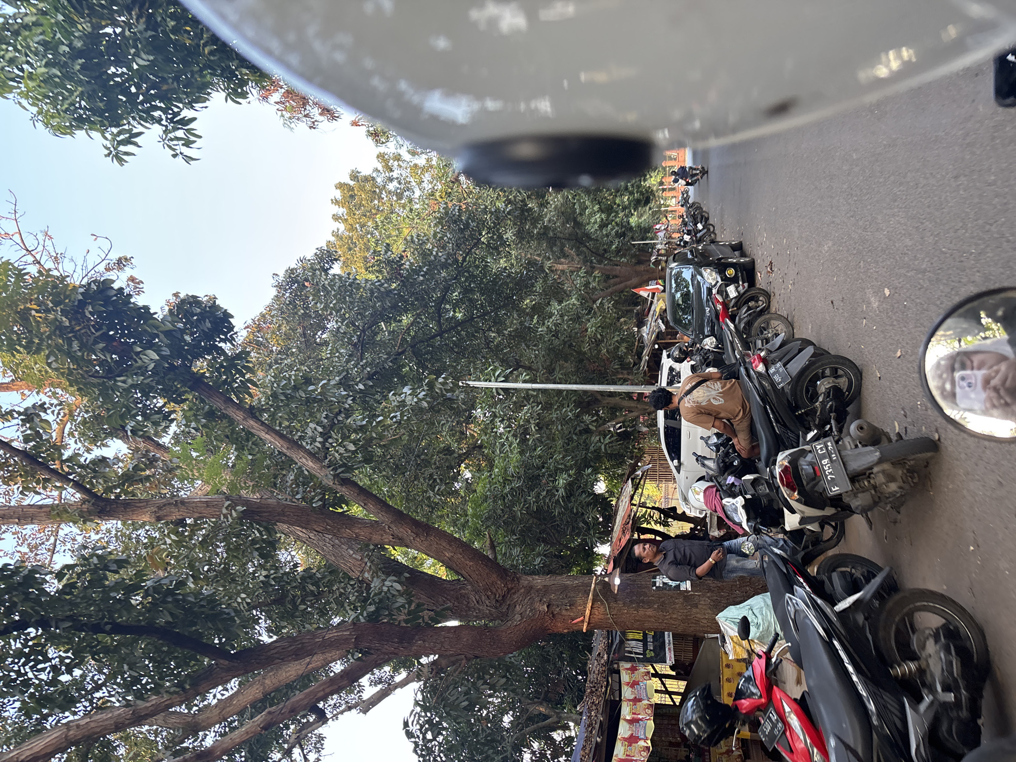A low-angle shot captures a street scene in Cirebon, Indonesia. In the foreground, the blurred white helmet of a motorcycle rider dominates the right side of the frame. To the left, several parked motorcycles are visible, including a red scooter and black scooters. A man in a dark shirt stands next to a large tree, facing a small stall with awnings. Beyond the parked motorcycles, cars and more motorcycles are seen on the asphalt road lined with trees. The sky is clear and blue. In the reflection of the motorcycle's rearview mirror, a person holding a smartphone can be seen. The image depicts a typical street in Cirebon, characterized by its bustling traffic and roadside stalls.