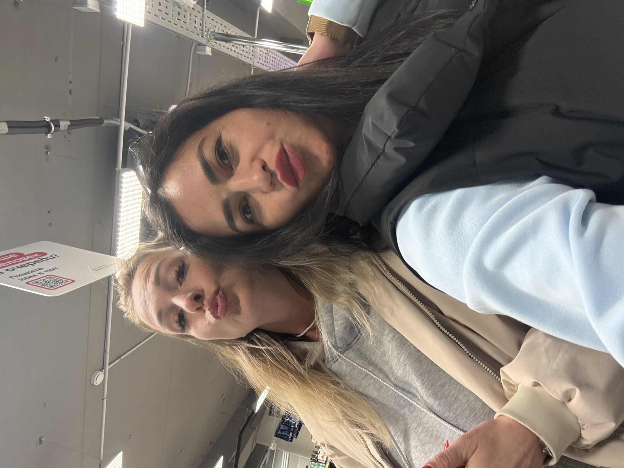 The image is a tilted close-up selfie of two women indoors, likely in Chaikovsky, Russia.
The woman in the foreground has long dark hair, dark eyes, and dark red lipstick, forming a "kissing" or "pouty" face. She is wearing a black puffer jacket over a light blue long-sleeved top. Her expression is playful.
Partially behind her, the second woman has long blonde hair and is also making a "kissing" face. She wears a grey hooded sweatshirt underneath a light beige jacket. Her expression mirrors the first woman's playful demeanor.
The background shows an indoor setting with an industrial-style ceiling, featuring exposed pipes, conduits, metal grating, and bright fluorescent lights. A white rectangular sign hangs from the ceiling. Visible Cyrillic text on the sign reads: "ЧЕЛО...В ОЧЕРЕДЬ?" (partially visible, likely "человек в очередь?" meaning "person in line?") and below it, "ПИШИТЕ НАМ В ЧАТ" ("WRITE TO US IN CHAT"). A QR code is also visible on the sign. The women appear to be engaged in a friendly, lighthearted moment.