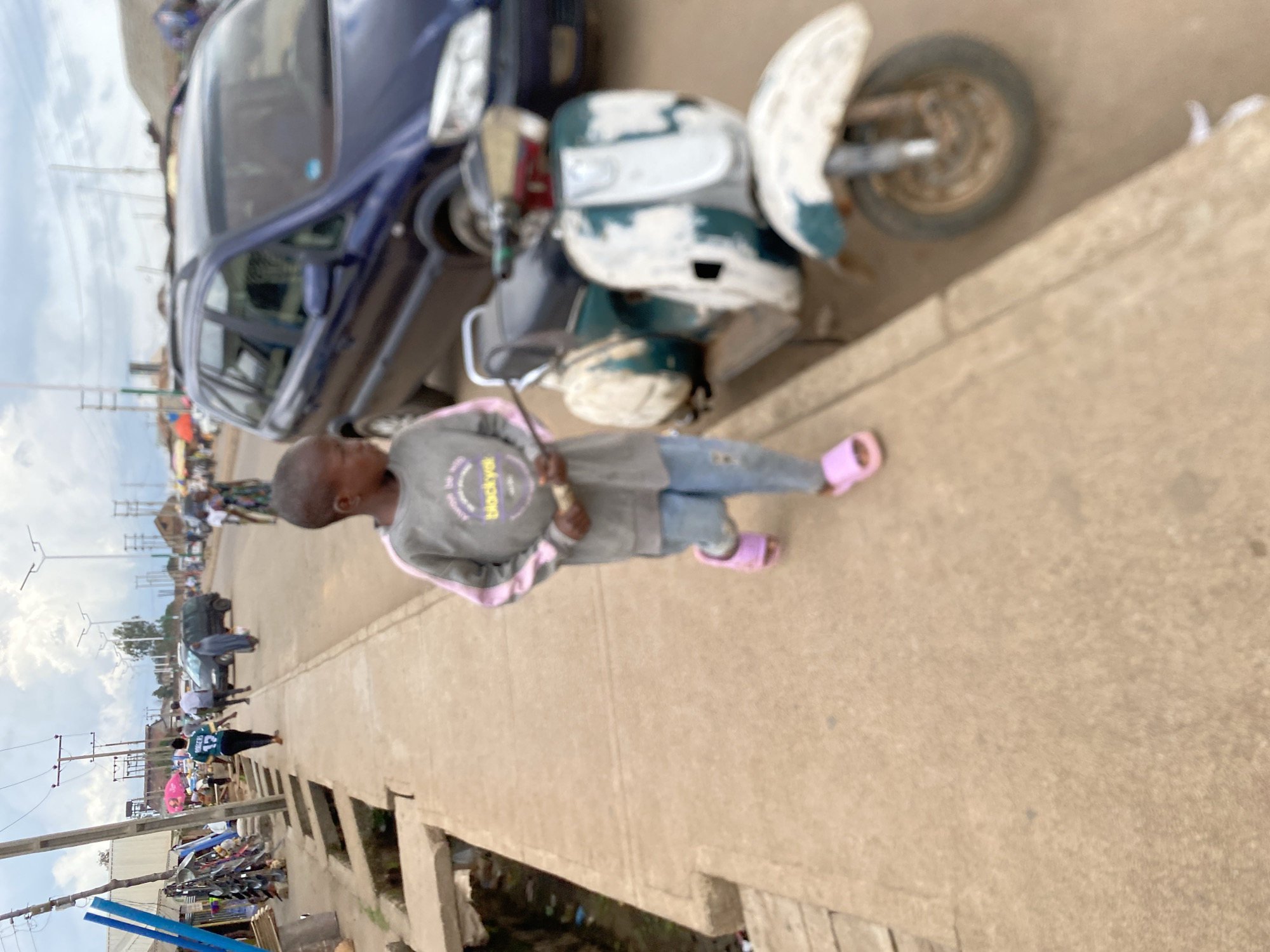 In Bukuru, Nigeria, a young boy walks along a concrete sidewalk adjacent to a street. He is wearing a gray sweatshirt with pink trim and light blue jeans, along with pink slide sandals. The boy is holding a thin stick in his hands. To his right, a green and white scooter is parked at an angle, its paint weathered and chipped. In the background, a blue minivan is visible on the street, along with other vehicles and pedestrians. The scene depicts a typical street environment with market stalls and various forms of transportation. The sky is overcast with scattered clouds.