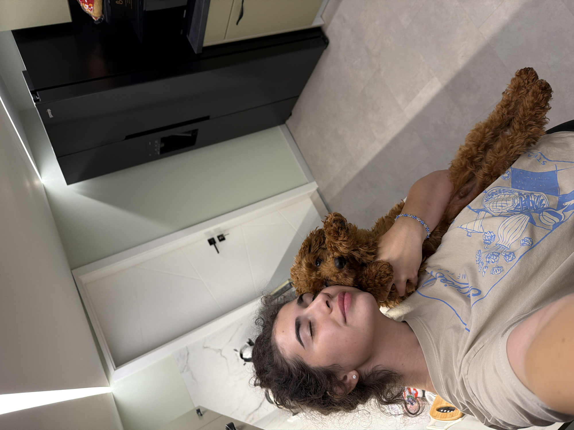 A young woman with curly brown hair is holding a small, brown dog. She is wearing a beige t-shirt with a blue graphic print. The dog is nestled against her cheek, and her eyes are closed. She is wearing a silver earring and a beaded bracelet on her wrist. The background appears to be a modern apartment kitchen, with a black refrigerator, a white door, and light-colored flooring. The scene is indoors. The image was taken in Sofia, Bulgaria.