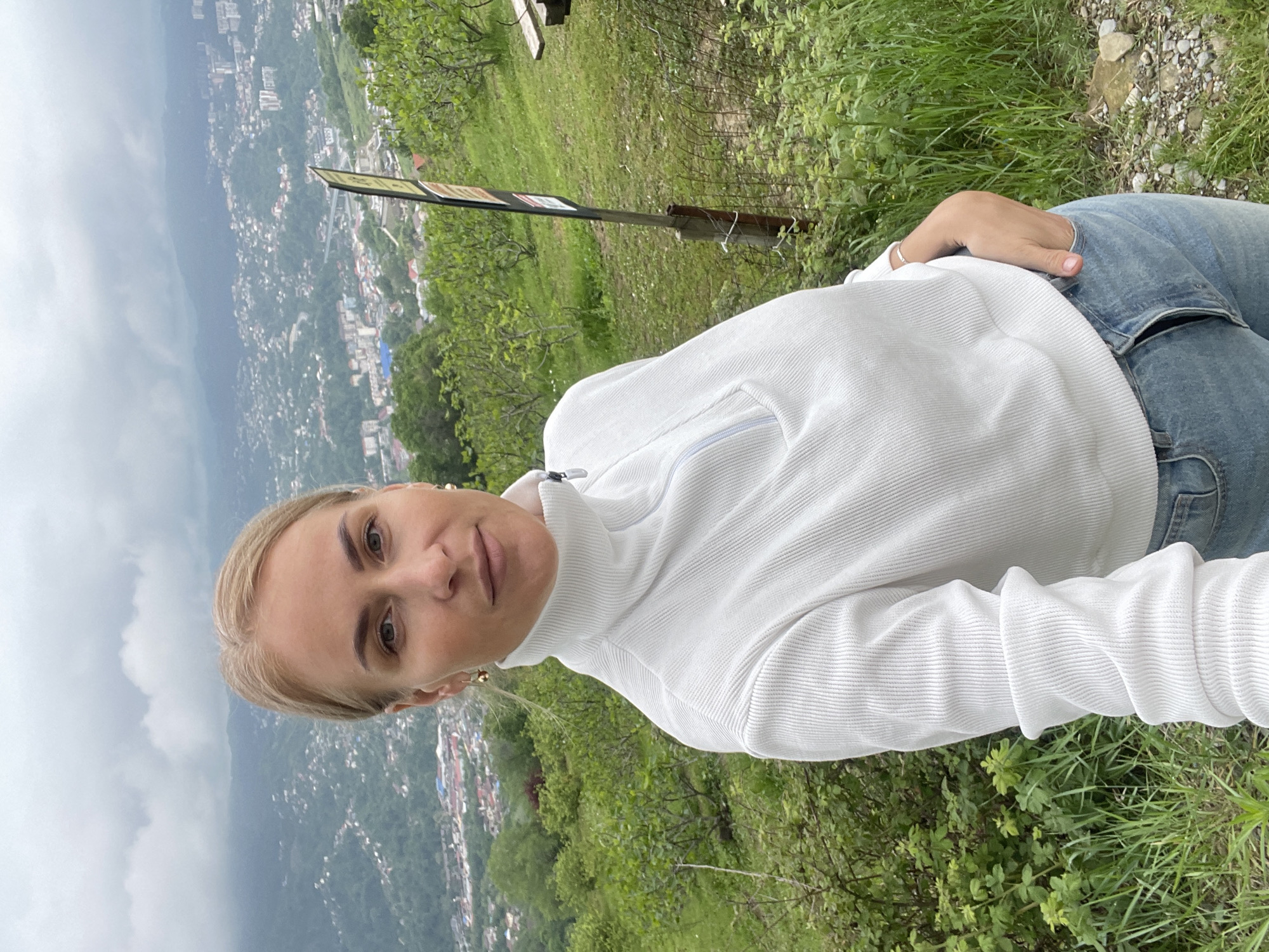 A woman is taking a selfie from a high vantage point overlooking a city, which is identified as Sochi, Russia, based on the urban layout and surrounding mountainous terrain. The woman is wearing a white turtleneck sweater and blue jeans. She stands on a grassy area with some foliage. Behind her, there is a partially visible sign and a metal barrier. The city sprawls across the landscape with buildings nestled within the hills and valleys, partially obscured by atmospheric haze. The sky is overcast.