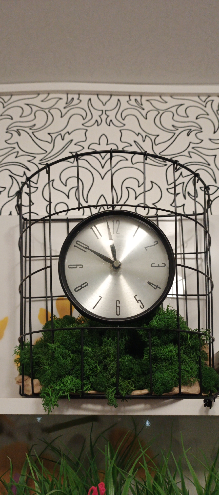 The image shows a clock within a black metal cage, filled with green moss. The clock face is silver with black numerals. Above the clock, a patterned wallpaper is visible. Below, artificial grass and a fish tank are partially visible. The picture was taken in Chaikovsky, Russia.