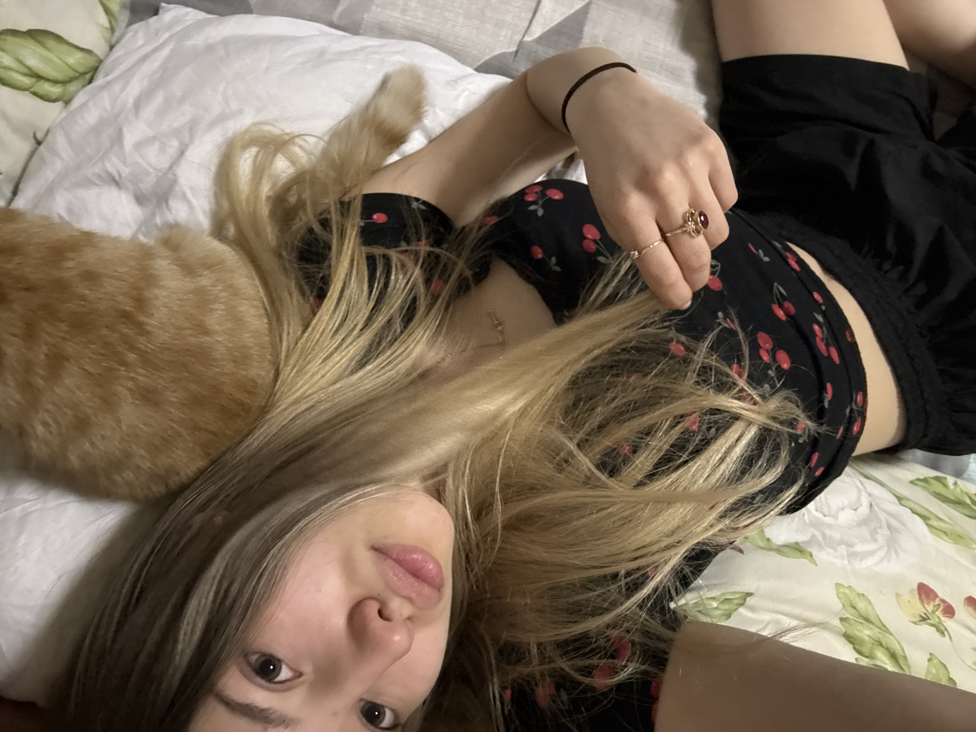 A woman lies on her back on a bed. An orange cat lies near her head on the left side. She wears a black dress with a cherry pattern. She has blonde hair and wears rings on her fingers. The bed has white and floral patterned sheets. The location is not discernible from the image.