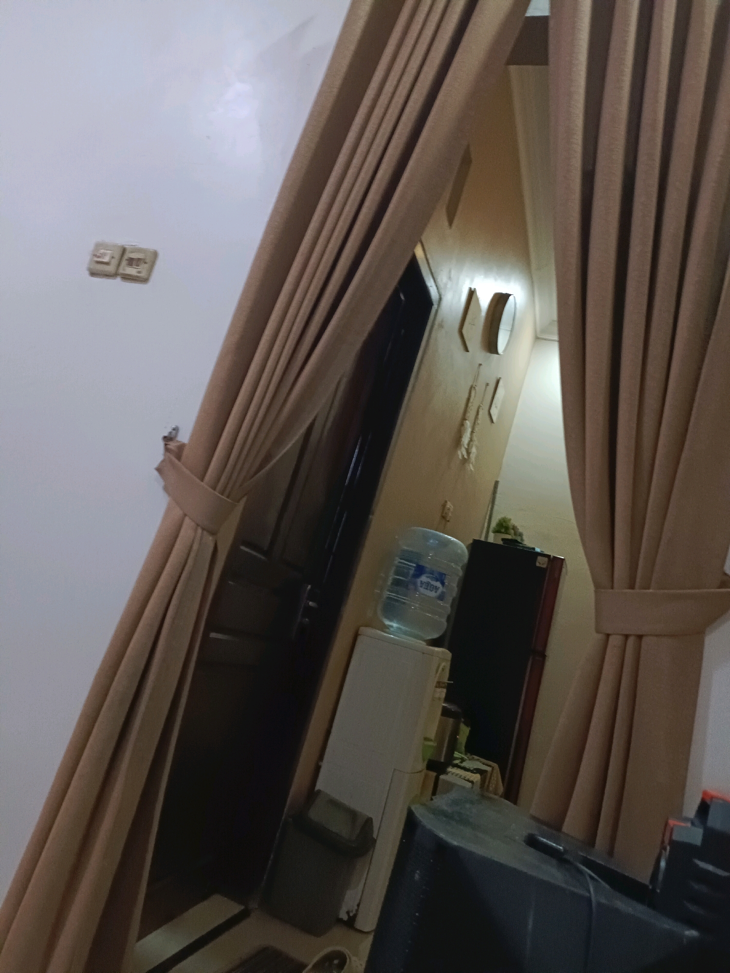 The image depicts an interior scene, likely a room or hallway, in Rancang Kencono, Indonesia. Heavy, beige curtains dominate the foreground, tied back with fabric bands, suggesting they are part of a window or doorway treatment. Through a gap in the curtains, a doorway is visible, leading into a dimly lit space. To the left of the doorway, a white water dispenser with a large blue water bottle is situated on a white appliance. Next to the water dispenser is a dark red refrigerator. On the wall above and to the right of the doorway, several framed objects and a round mirror are mounted. The wall surface has a textured finish. In the lower right of the image, a dark object, possibly a television or monitor, is visible. The overall lighting is low, creating shadows and a somewhat muted atmosphere.