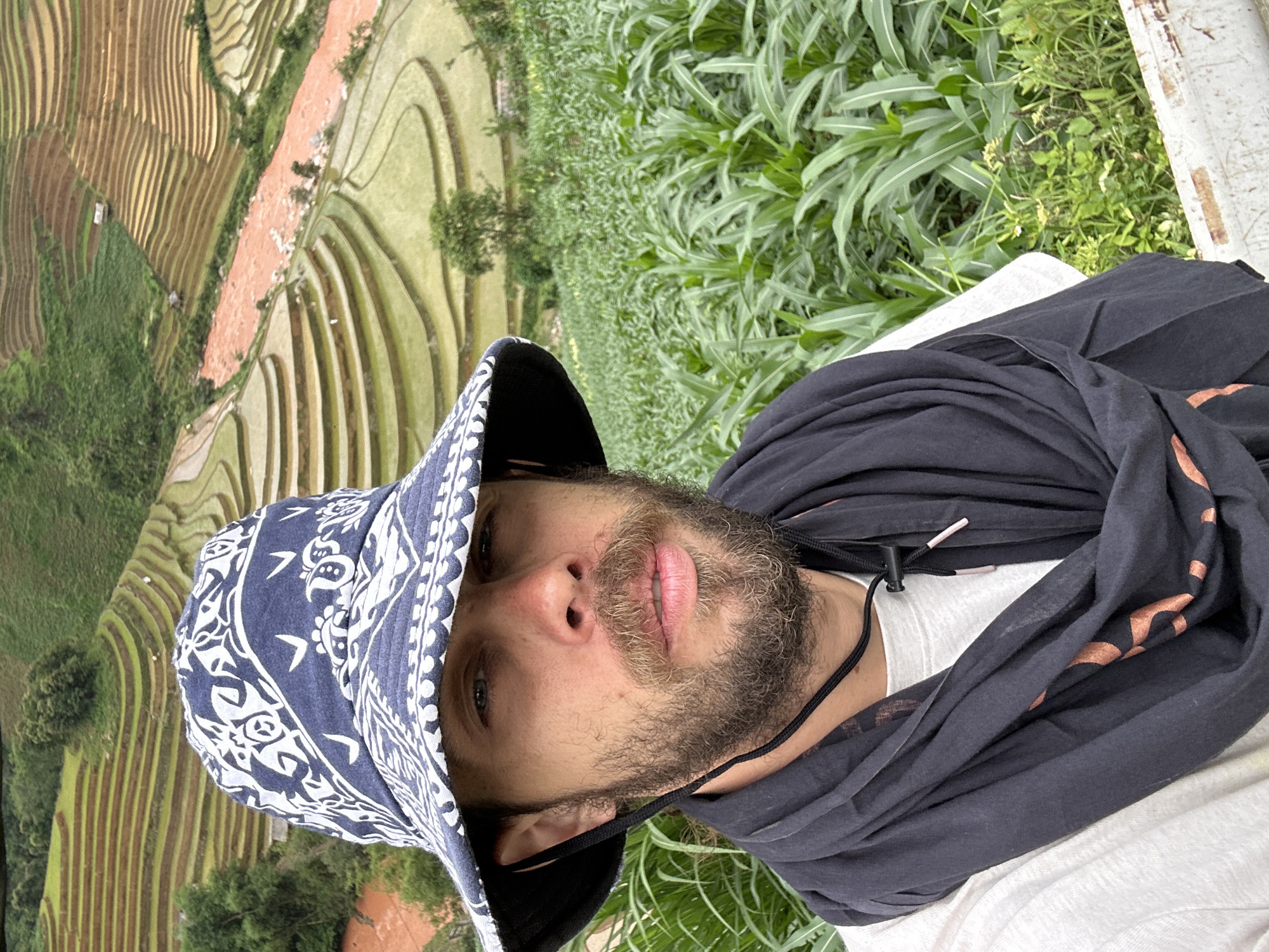 A male subject with a beard and mustache, wearing a dark blue patterned bucket hat, a black scarf, and a white t-shirt, is depicted from the chest up, looking directly at the camera. The setting is Mù Cang Chải, Vietnam, characterized by extensive agricultural terraces. In the background, vibrant green rice terraces and brown, recently tilled or fallow terraces are visible on undulating terrain. A distinct reddish-orange river flows through the terraced landscape. The immediate foreground to the subject's right features a field of tall green crops, likely corn. Sparse trees and vegetation cover the hillsides, and small structures are present within the distant terraces.