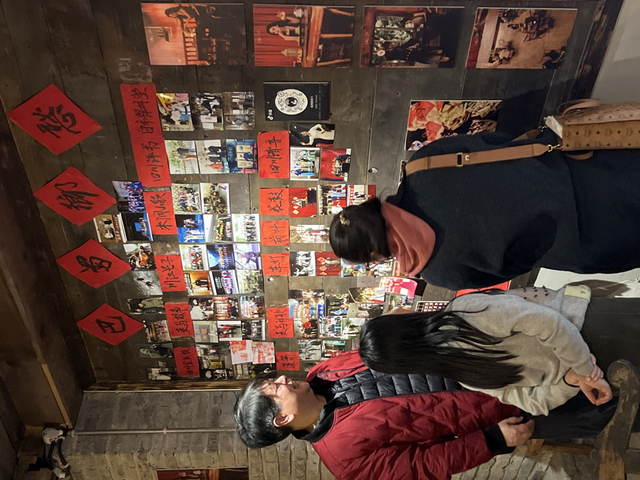 An indoor scene, identified as being in Yuzhong District, China, captures three individuals observing a wall densely covered with photographs and red calligraphic decorations.
An adult male stands on the left, facing slightly right, wearing a red quilted jacket over a dark underlayer. He possesses dark hair and exhibits a slight smile while looking towards the other two individuals.
A young female, likely a child or pre-teen, is positioned in the center, viewed from behind. She has long, straight dark hair and wears a light grey long-sleeved top over dark trousers or a skirt. Her hands are clasped behind her back as she gazes at the wall display.
To the right of the young female, an adult female is also observed from behind. Her dark hair is styled away from her face, and she wears a dark top layered with an orange/rust-colored cowl-neck garment. A light brown shoulder bag with a distinct patterned monogram hangs on her right side. She is focused intently on the wall.
The primary feature of the environment is a dark-stained wooden plank wall serving as a display surface. Four red diamond-shaped papers with black Chinese calligraphy, including characters such as '巴' (Ba), '罗' (Luo), '御' (Yu), and '慈' (Ci), are affixed near the top of the wall. Numerous smaller rectangular red papers with various Chinese phrases, such as '四川评书 白蛇传学堂' (Sichuan Pingshu, White Snake Legend Classroom), '四川清音' (Sichuan Qingyin), and '川江号子' (Chuanjiang Haozi), are interspersed among a multitude of photographs.
The photographs, varying in size, depict diverse cultural and performance-related themes, including group portraits, individuals in traditional costumes, stage performances, and interior settings. A column of larger, vertically oriented photographs on the far right showcases stylized scenes, one featuring an individual at a table with tea, and another an overhead view of a room with seated figures. One photograph includes a visible logo associated with '抖音' (Douyin/TikTok).
To the left of the individuals, a segment of a textured brick or stone wall is visible. The scene is illuminated by warm, even lighting. The three individuals are engaged in silent observation of the wall's comprehensive cultural display.