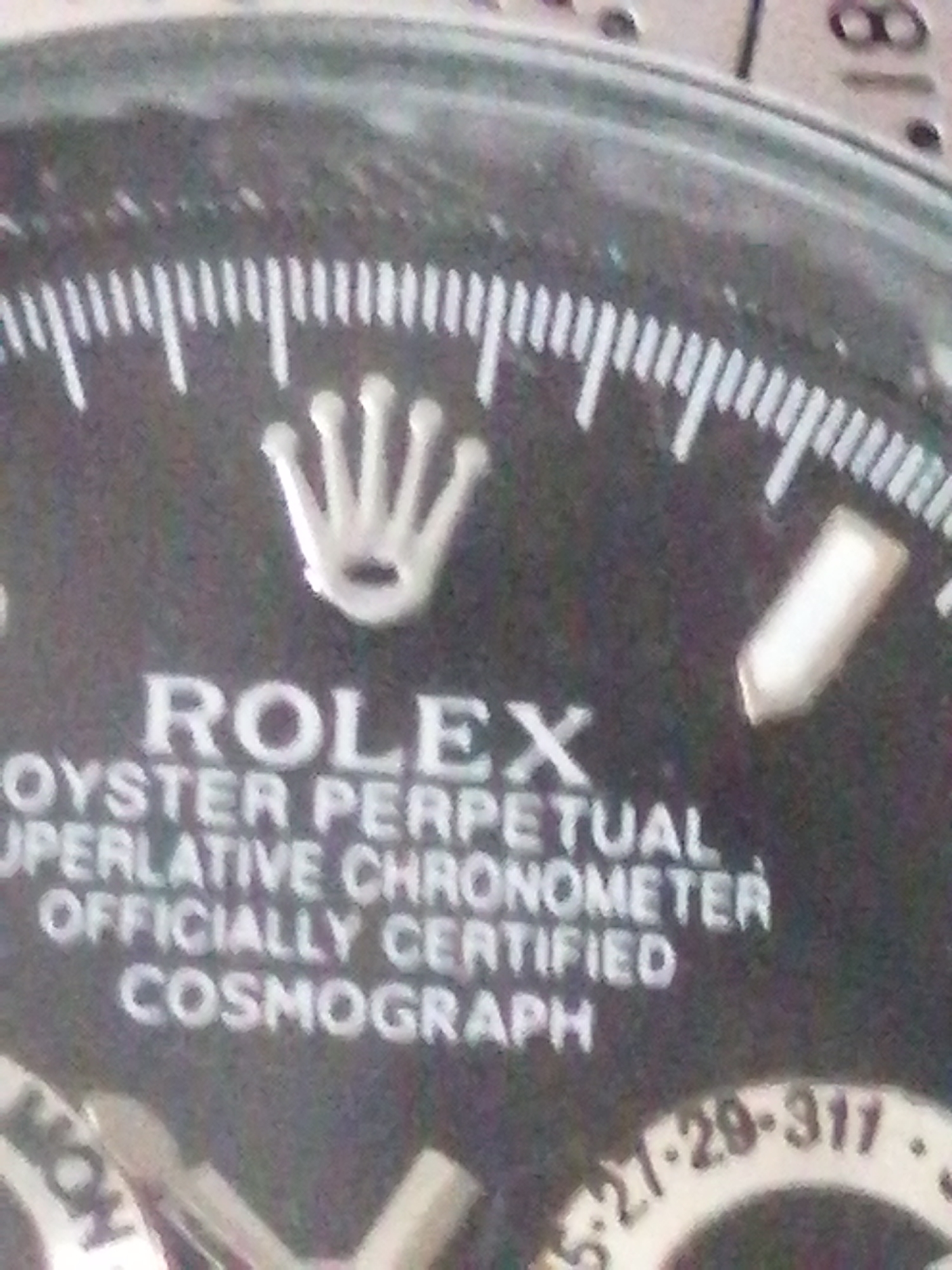 The image shows a close-up of a Rolex watch face. The watch has a black dial with white markings. The Rolex crown logo is prominently displayed at the 12 o'clock position. Below the crown, the text "ROLEX" is written in large white letters, followed by "OYSTER PERPETUAL", "SUPERLATIVE CHRONOMETER", "OFFICIALLY CERTIFIED", and "COSMOGRAPH". There are also white hour markers and minute markers around the outer edge of the dial. A white hour hand and minute hand are visible, along with a thin white second hand. A tachymeter scale is visible around the bezel, with numbers such as "27-29-317" partially readable. The overall impression is of a high-quality, classic timepiece. The image is from an unknown location in an unknown city.