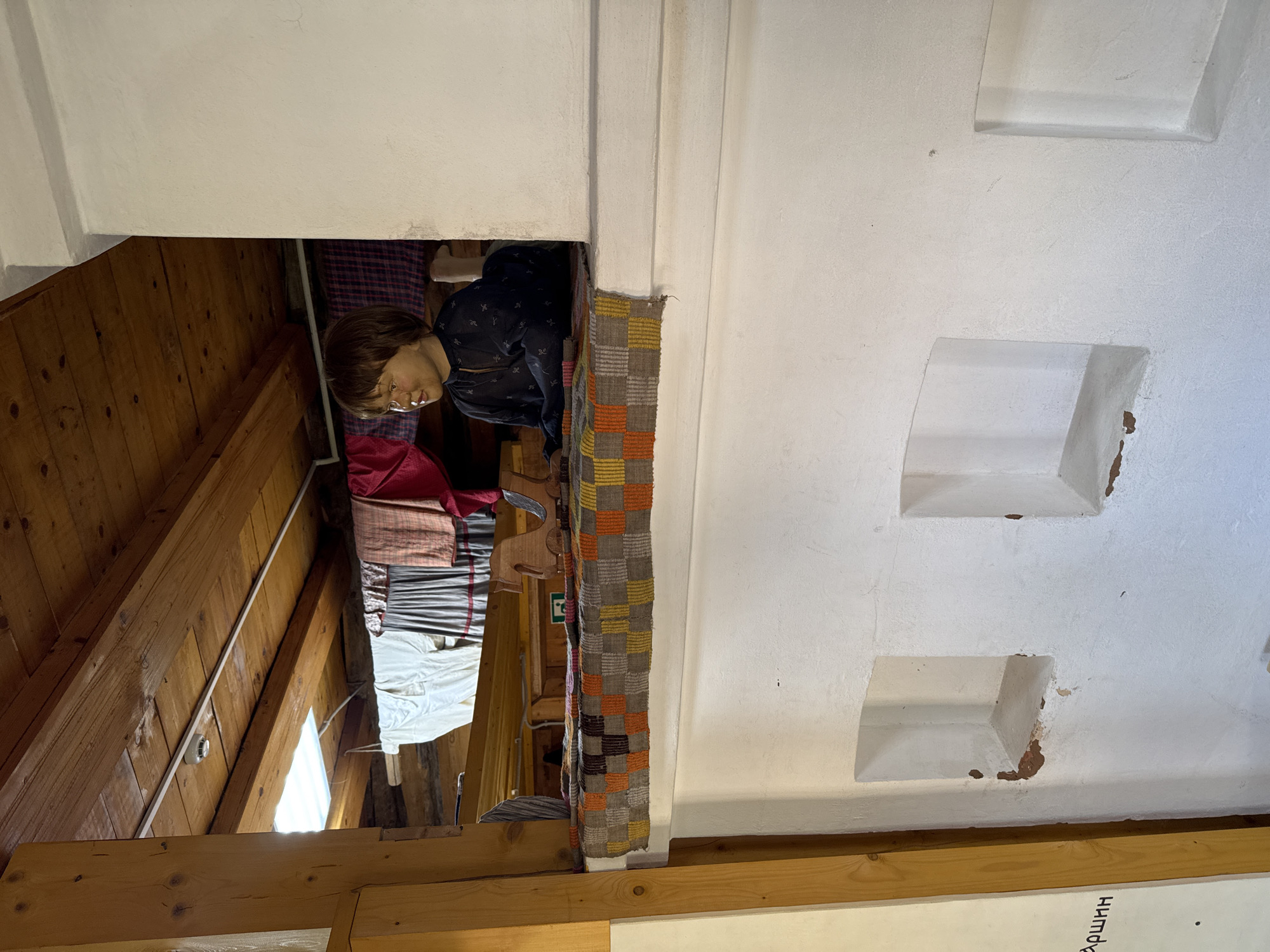 The image depicts the interior of the Peter Tchaikovsky Museum in Votkinsk, Russia. The view is framed by a doorway. Inside the room to the left, a mannequin dressed in period clothing sits at a table. Multiple layers of textiles or bedding are visible on the table. The walls of the room are made of wooden planks. Outside the doorway, the wall is painted white and features recessed rectangular niches. Wooden beams are visible at the bottom of the frame, with some writing in a Cyrillic script below.