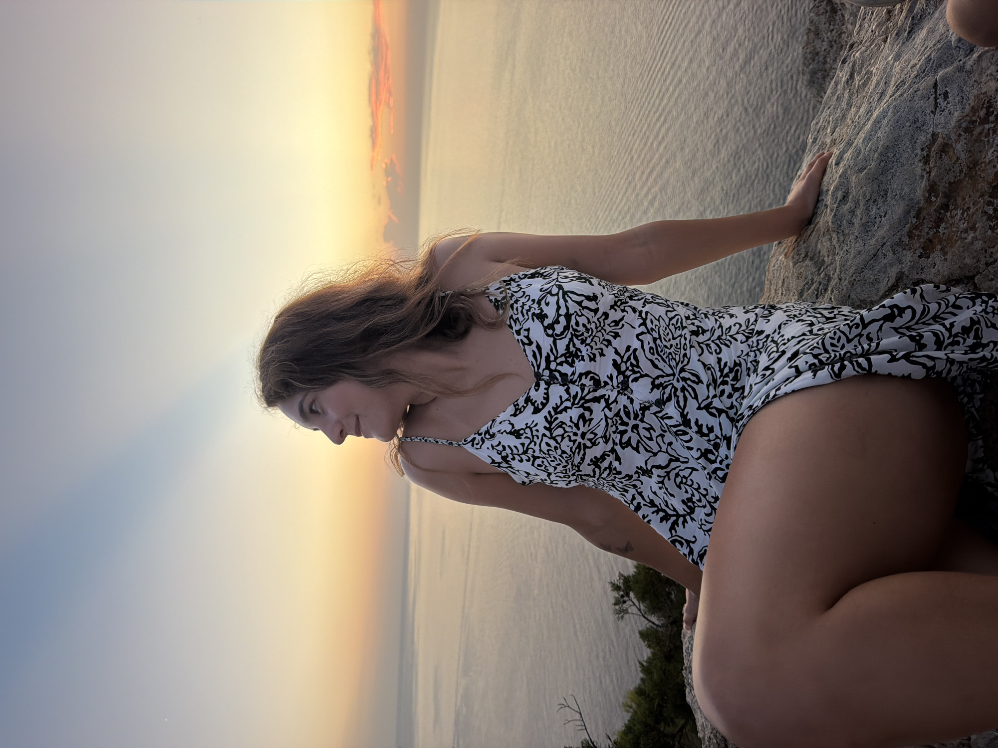 A young woman with long, wavy brown hair is seated on a rocky outcrop, looking out over a calm body of water. The setting appears to be a coastal area, possibly in Sant Antoni de Portmany, Spain, given the time of day and the characteristic serene seascape. She is wearing a black and white patterned sundress with thin straps. Her legs are crossed, and her left arm rests on the rock beside her. The sky is painted with soft hues of orange, yellow, and blue, indicative of either a sunrise or sunset. The water reflects the sky's colors and shows subtle ripples. Some sparse vegetation can be seen on the left side of the frame, growing from the rocky terrain. The overall mood of the image is peaceful and contemplative.