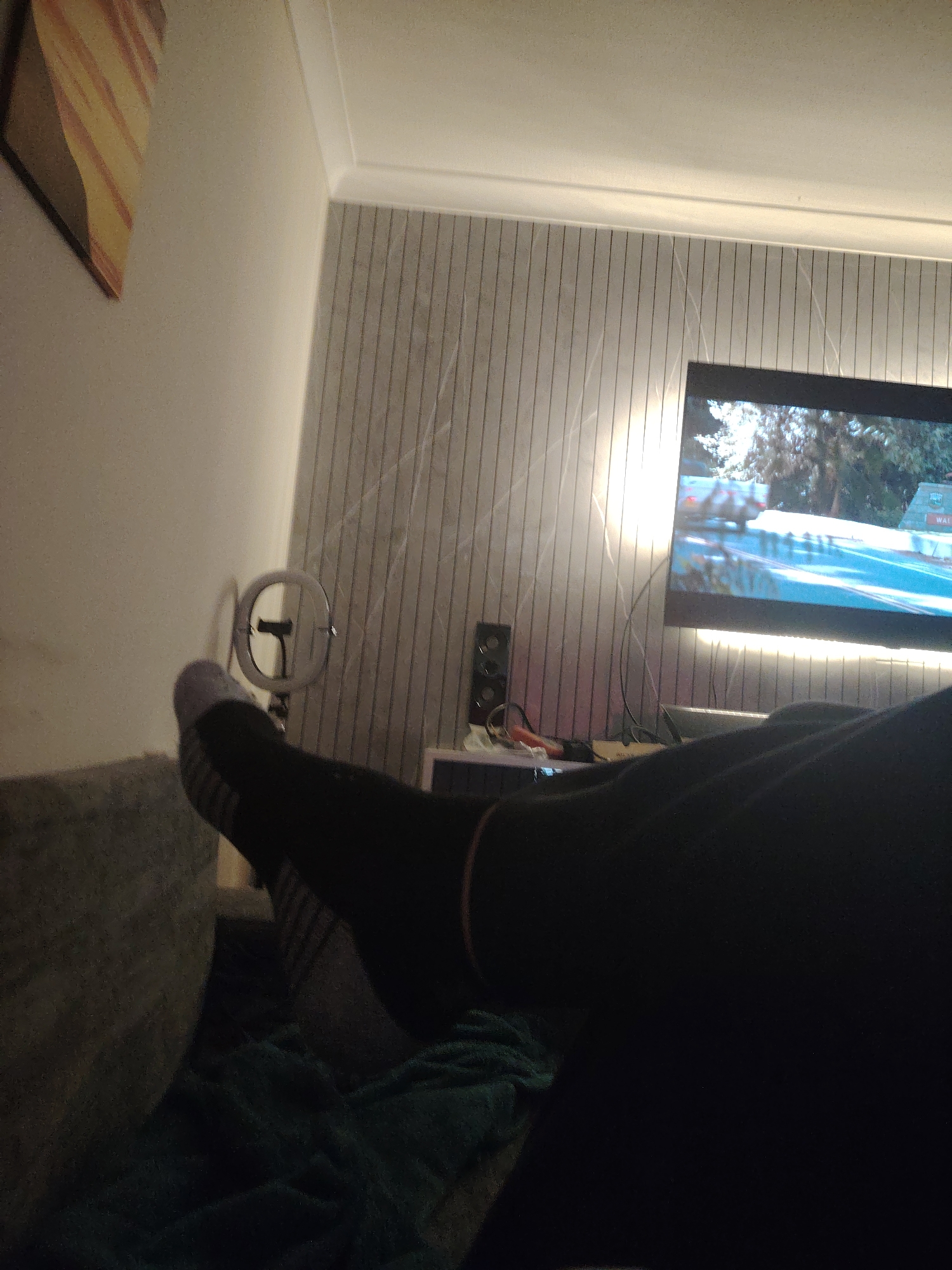 The image shows a person's legs and feet, clad in black leggings and grey and white striped socks, propped up on a grey sofa. A dark green blanket is visible beneath the legs. To the right, a television screen displays a video game scene with a car on a road. A black speaker is positioned below the TV, next to some electronic devices and cables. In the background, a wall is decorated with vertical lines, and a light fixture runs along the top. A round, metallic ring light is visible on a stand between the sofa and the wall. On the left side of the image, a portion of a wall-mounted artwork with diagonal stripes of yellow, orange, and brown is partially visible. The scene appears to be an indoor living space.