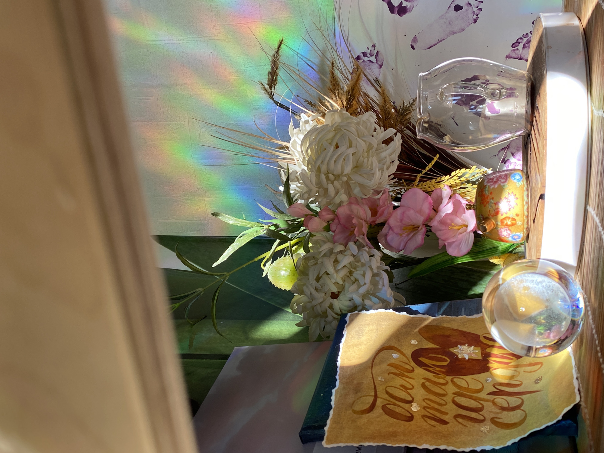 The image depicts a still life arrangement. In the foreground, there's a sheet of parchment with Cyrillic calligraphy in brown ink, scattered with small silver leaf accents. A clear glass paperweight sits atop this parchment. Behind and to the left of the parchment are floral arrangements featuring white chrysanthemums, pink orchids, and green foliage. A decorative gold cylinder with floral patterns is visible, alongside a clear glass vessel on a wooden surface. The backdrop is a wall, illuminated by a rainbow prism effect, with faint purple footprints adorning its surface. A partially visible light-colored curtain is on the left side of the frame. The image does not contain information that identifies the location as Chaikovsky, Russia.