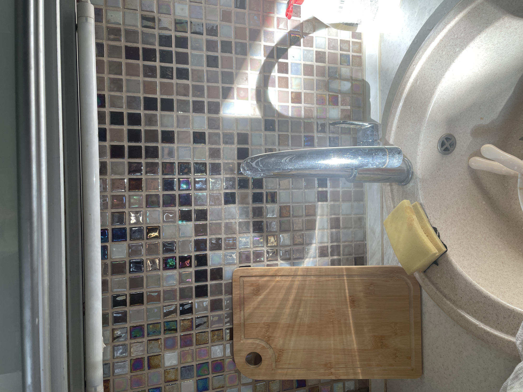 The image shows a kitchen sink area with a tiled backsplash. In the foreground, a beige sink basin is visible, with a chrome faucet extending upwards. A yellow sponge rests on the edge of the sink. To the left of the sink, a wooden cutting board with a circular handle hole is placed on the countertop. The backsplash consists of small, square tiles in various shades of white, grey, black, and iridescent colors, reflecting the sunlight. The overall environment appears to be a domestic kitchen setting. There is no specific indication that this location is in Kaliningrad, Russia.