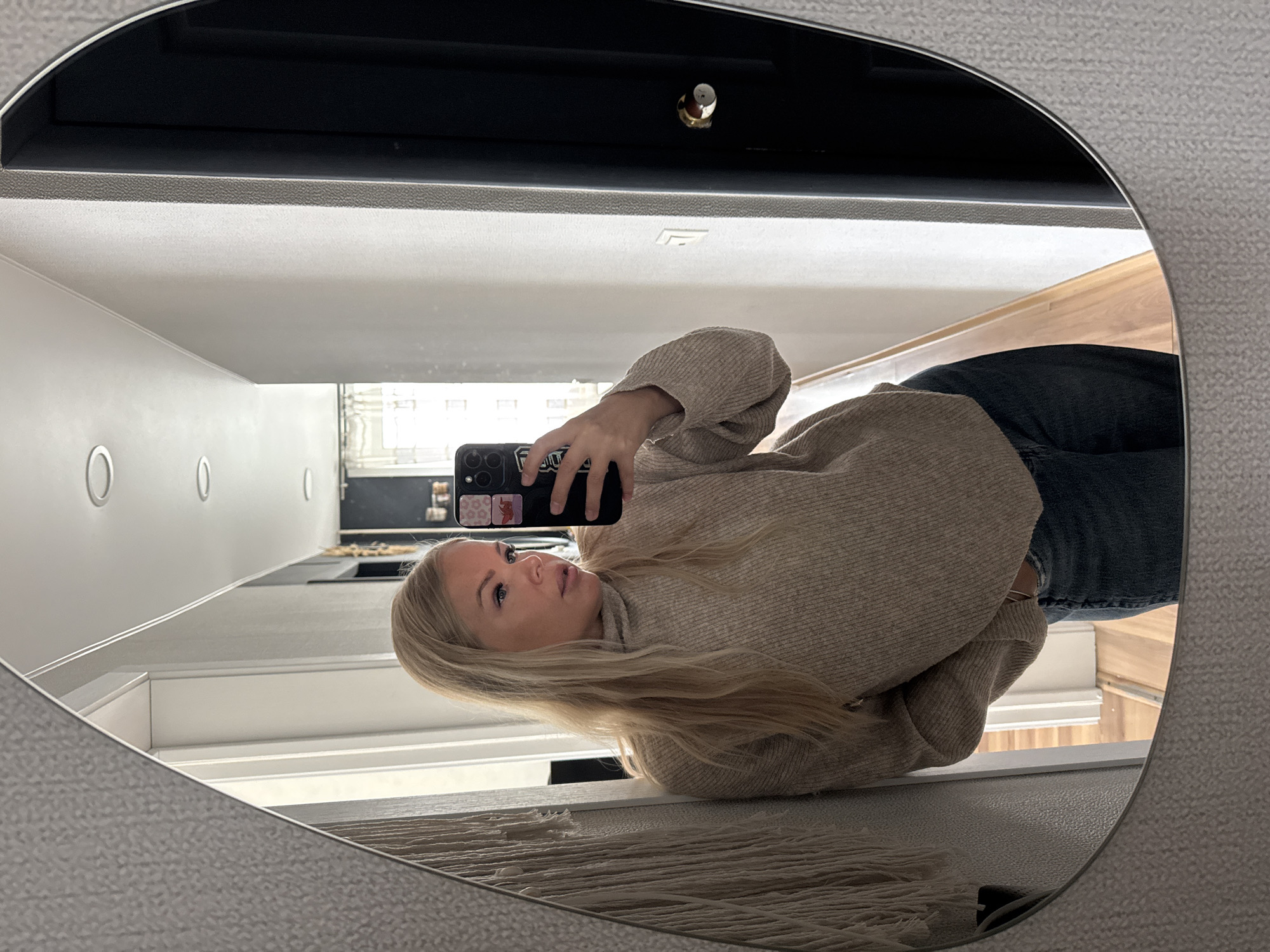 A blonde woman takes a mirror selfie indoors, reflected in an irregularly shaped mirror with a light gray, textured frame. She is wearing a light beige ribbed turtleneck sweater and dark blue jeans, holding a dark smartphone with a multi-lens camera and decorative stickers, one featuring leopard print and another possibly bearing the text "FOLK". Her long hair falls over her shoulder, and she looks subtly upwards towards the phone.
The reflections in the mirror reveal the surrounding environment. Above the woman's head, a dark horizontal surface, possibly a shelf or cabinet, with a small metallic knob or handle, is visible. Below this, a lighter textured strip leads to a white panel featuring multiple circular indentations, suggesting cabinet or drawer handles. To the lower right, a warm-toned wooden floor can be seen, along with a light-colored, fringed textile item, possibly a rug or throw. The background glimpsed through the reflection indicates an interior space, likely a modern room or hallway, well-lit, suggesting daytime.