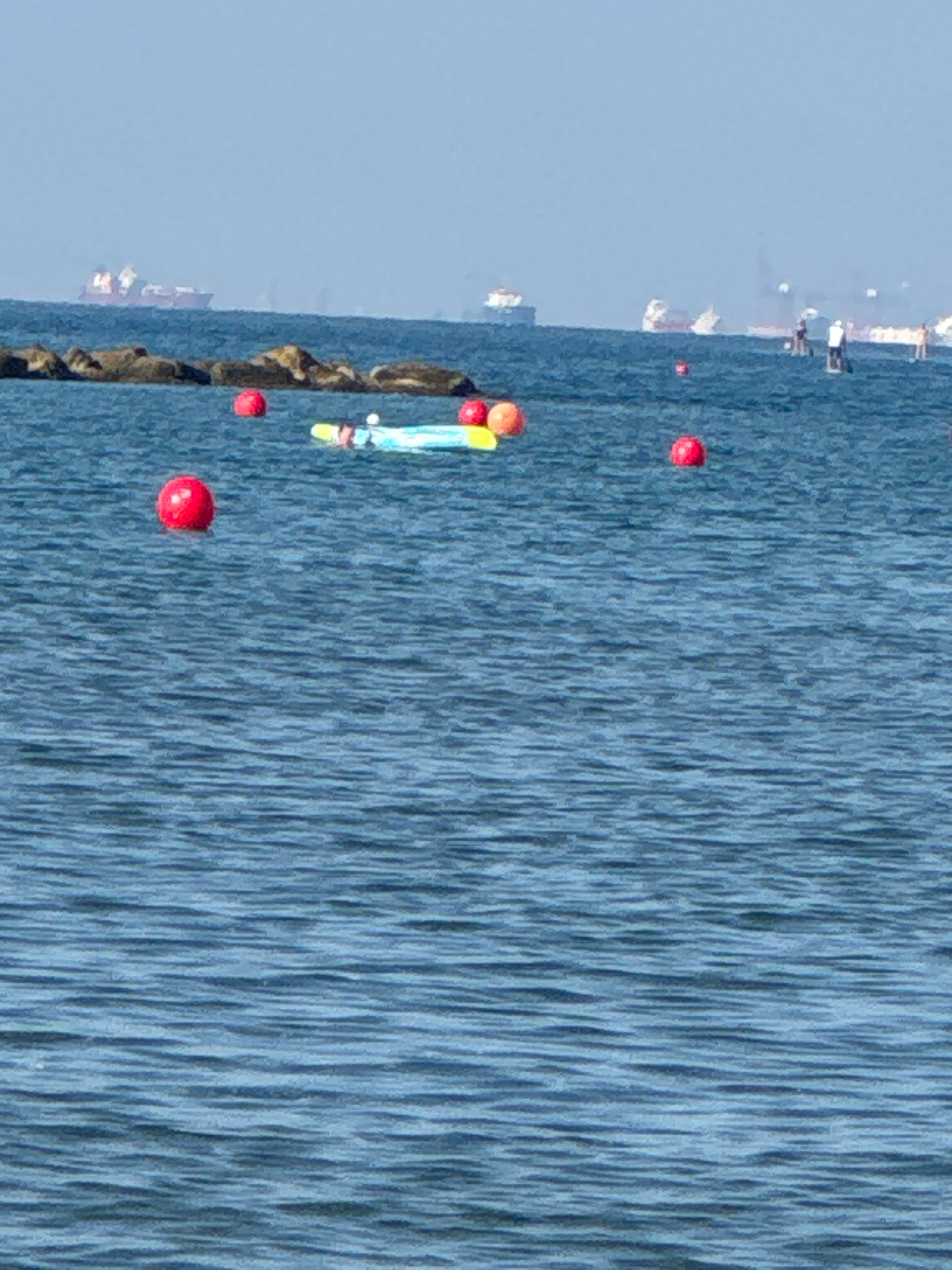 The image depicts a body of water, likely the sea, with several objects floating on the surface. In the foreground, there are several red spherical buoys. Further back, a person is visible on a paddleboard, and another person appears to be sitting on a yellow and blue inflatable raft. In the distance, several large cargo ships are visible on the horizon, with one ship prominently displaying a red hull. The water is a deep blue with visible ripples. The sky is a pale blue and hazy. This scene is consistent with the coastal waters of Agios Tychona Community, Cyprus, a region known for its shipping activity and recreational water use.