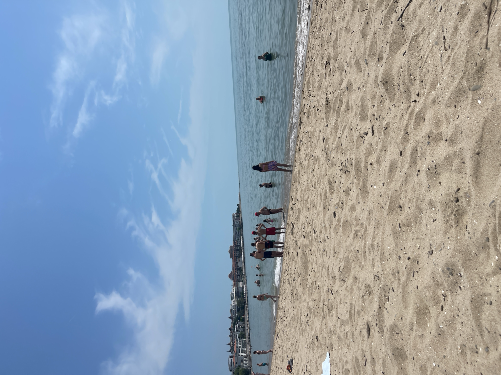 The image captures a vibrant beach scene in Sveti Vlas, Bulgaria, under a clear, sunny sky with scattered white clouds. The picture is vertically oriented.
In the foreground, a wide expanse of light brown sand stretches across the right side of the frame, showing ripples, footprints, and small pebbles. The mid-ground features a calm body of water, likely the Black Sea, where numerous people are engaging in various activities. Several individuals are seen swimming or wading deeper in the water. Closer to the shore, groups of people, including adults and possibly some children, are standing or walking along the water's edge, enjoying the warm weather. They are all dressed in swimwear.
Along the distant coastline in the background, a series of multi-story buildings are visible, indicating a developed resort town or residential area lining the shore. The time of day appears to be midday, given the bright illumination and distinct shadows. The overall atmosphere is one of leisure and enjoyment on a pleasant summer day. No visible text is present in the image.