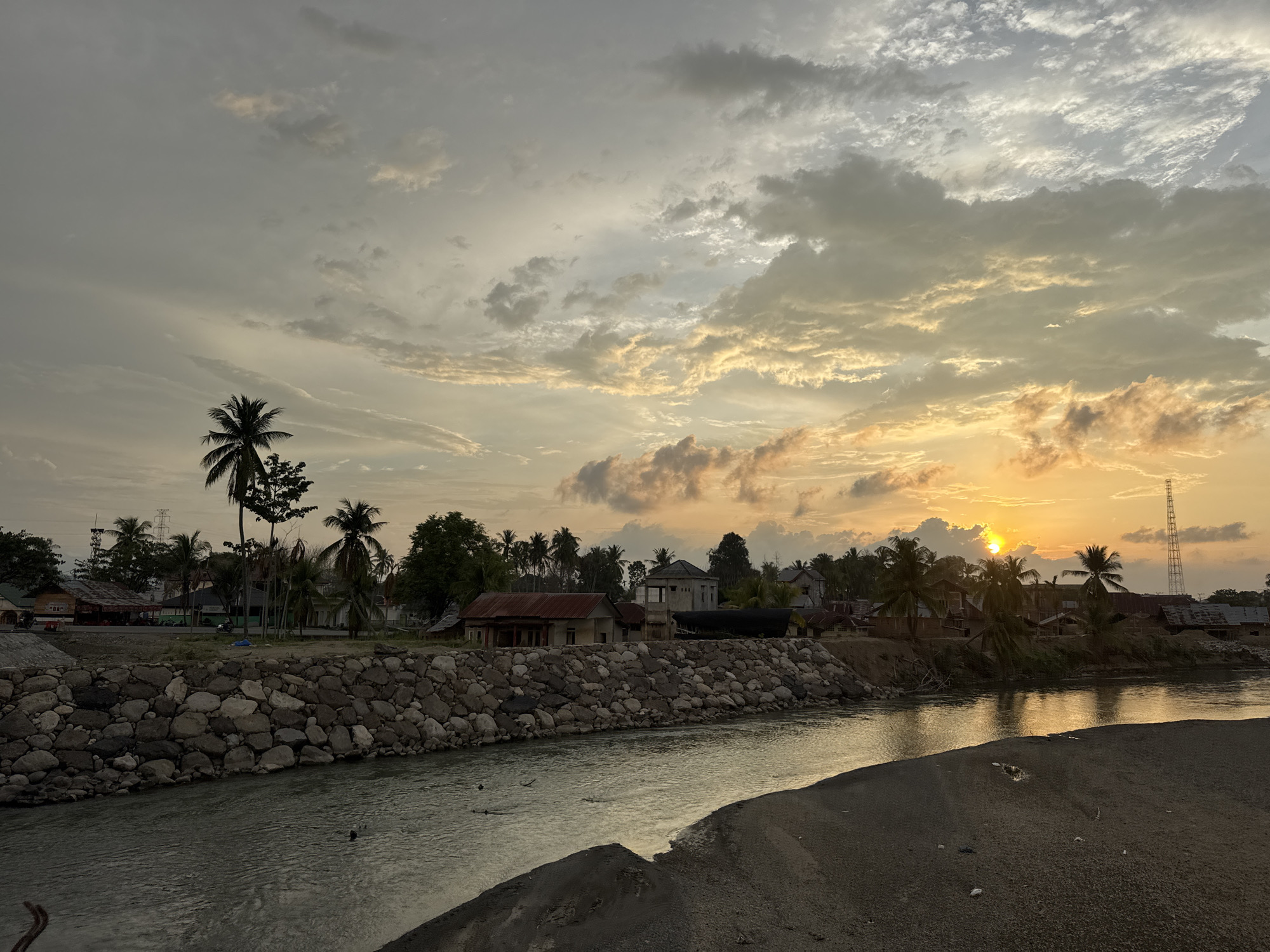 This media file depicts a serene sunset scene in Meunasah Pupu, Indonesia. The setting is a riverbank lined with a stone wall, with a calm river flowing in the foreground. Beyond the wall, a small village is visible, characterized by houses with red and brown roofs, palm trees, and a few other trees. A transmission tower can be seen in the distance on the right side of the frame.
The time of day is clearly sunset, as indicated by the golden hues in the sky and the low angle of the sun, which is partially obscured by clouds. The sky is filled with a mix of dramatic clouds, some dark and heavy, others illuminated by the setting sun, creating a picturesque and slightly moody atmosphere. The weather appears to be pleasant, with the sun breaking through the clouds.
There are no people visible in the scene, but the presence of houses and a road suggests human habitation and activity in the area. The overall emotion conveyed by the image is peaceful and tranquil, capturing the beauty of a tropical sunset. No visible text is present in the image.