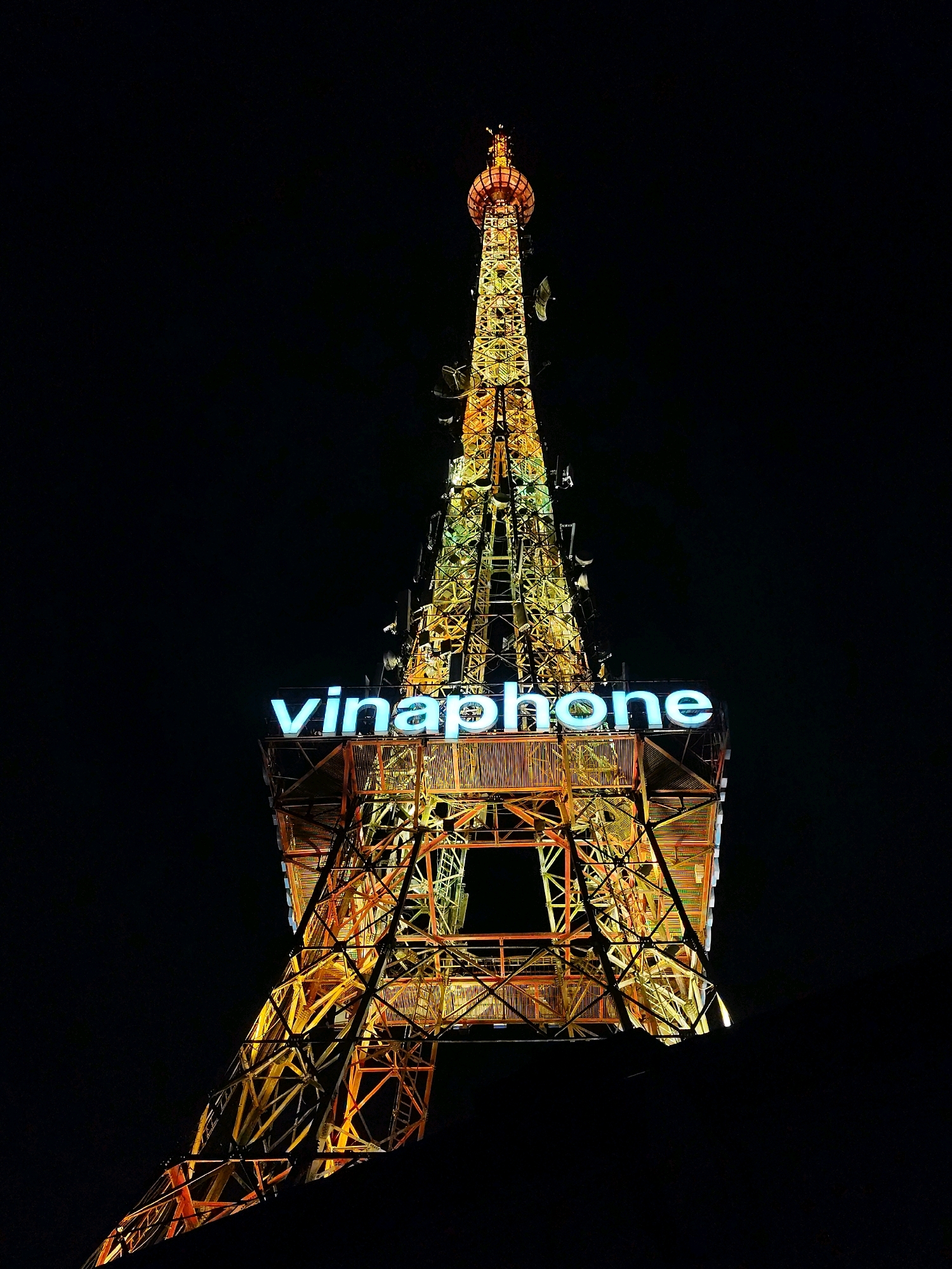 The image shows the Da Lat Communications Tower, also known as the "Vinaphone Tower," in Da Lat, Vietnam, at night. The tower is illuminated with yellow and orange lights, giving it a warm glow against the dark sky. A large "vinaphone" logo, with blue neon letters, is prominently displayed on the tower. The tower's intricate metal lattice structure is visible, and several antennas are mounted on its sides. The overall scene is one of an illuminated, modern structure standing tall in a dark, urban environment.