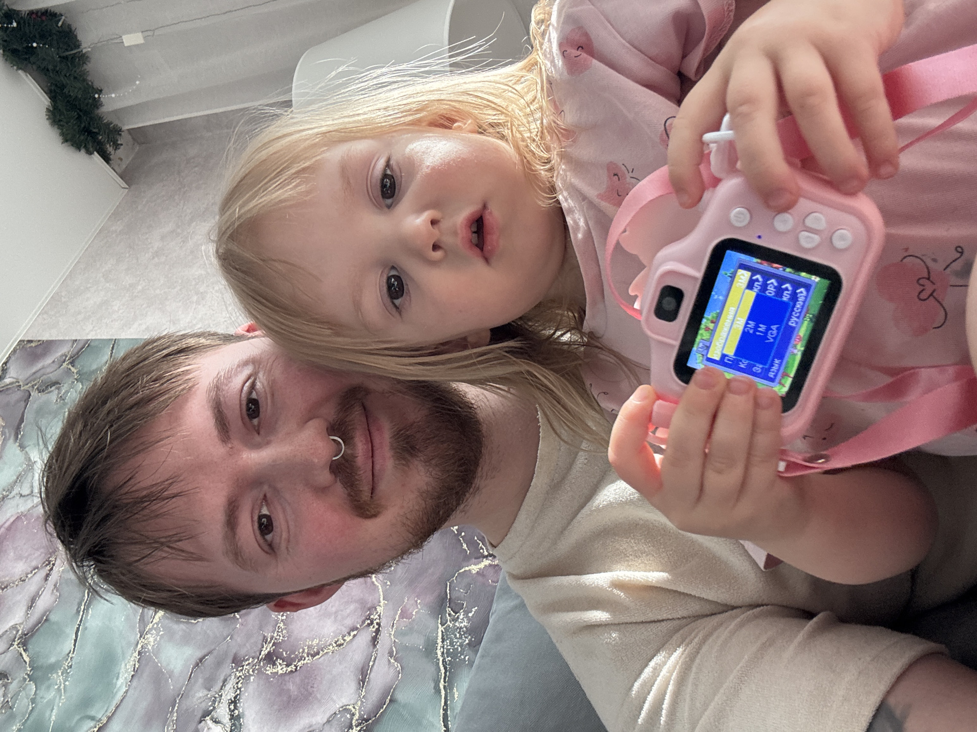This tilted, close-up image captures a candid moment between a man and a young child indoors in Chaikovsky, Russia. The adult man, with light brown hair, a beard, and a septum piercing, occupies the left side of the frame, looking towards the camera with a slight smile. He is wearing a light-colored long-sleeved top.
Resting on his shoulder or chest, positioned to the right and slightly above him, is a young child, likely a girl, with blonde hair and light-colored eyes. She wears a pink long-sleeved top with light patterns and looks attentively towards the camera with her mouth slightly open.
The child is holding a small, pink digital camera with a pink strap. The camera's screen is active, displaying a menu interface. Clearly visible text on the screen includes "Русский" (Russian), indicating the language setting, and resolution options such as "3M," "2M," "1M," and "VGA." The word "изображения" (images) is also partially visible.
The background reveals an indoor setting, likely a home, with a light-colored wall and ceiling visible on the left. A green Christmas garland or decoration is placed on a shelf in the upper left corner. Below the man, a soft surface, possibly a blanket or cushion, features an abstract pattern of swirling light purple, blue, and metallic gold/white. The bright, diffused lighting suggests it is daytime. The image conveys a warm, domestic scene.