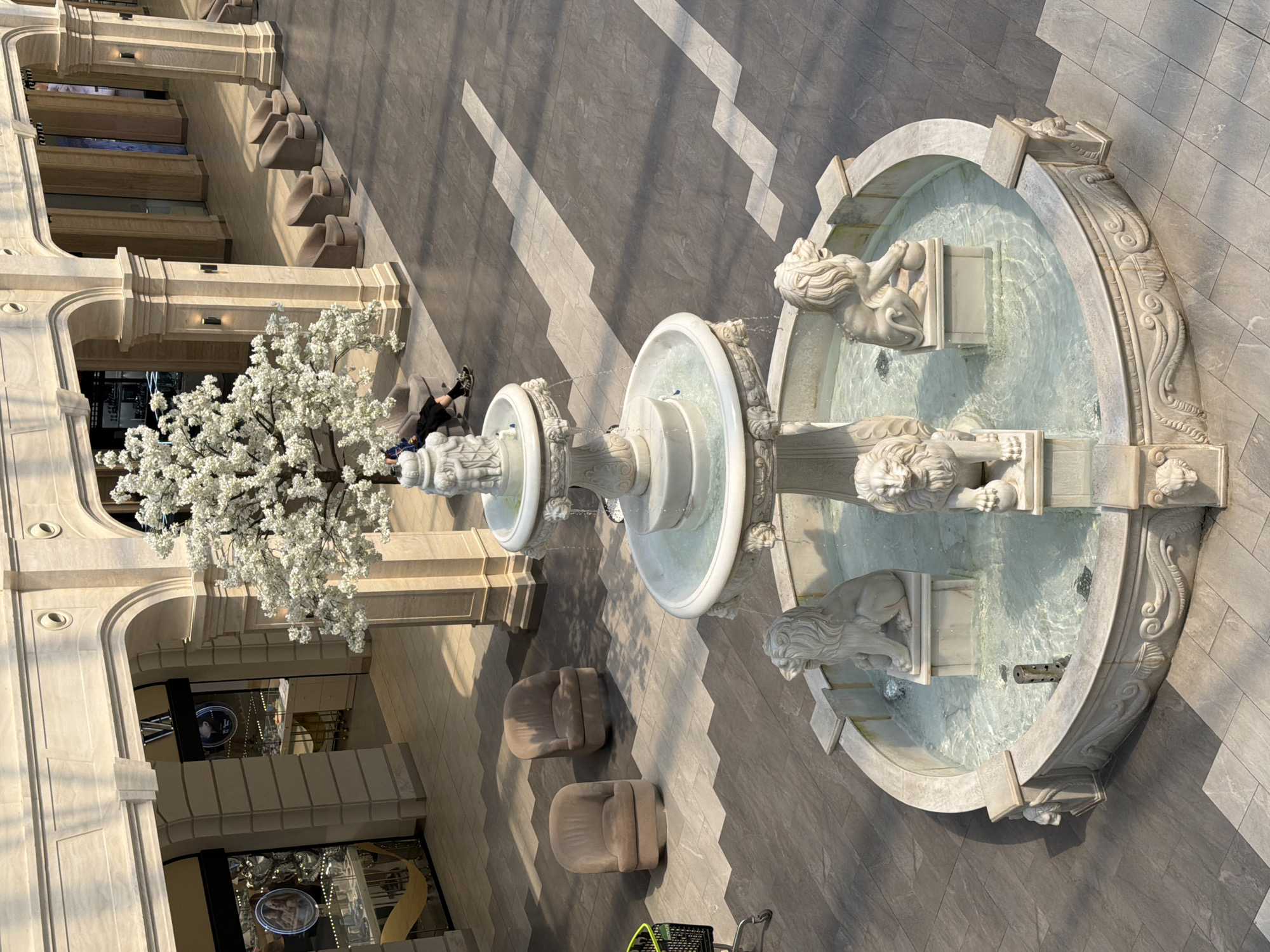 This is a photograph of a multi-tiered ornamental fountain with lion sculptures in the foreground, located in a modern indoor shopping mall or atrium. The fountain has a white stone basin with intricate carvings, and the water is clear with visible ripples. Four lion statues are positioned around the basin, two of which are visible in the lower part of the frame. Above the fountain, a large artificial tree with white blossoms is prominent, and behind it, a person is visible sitting on a chair. The surrounding environment features cream-colored architectural columns and arches, with shops visible in the background through glass storefronts. The floor is tiled in a pattern of dark grey and light grey squares. The scene is well-lit, suggesting an indoor space. The location in Kyiv, Ukraine is not identifiable from the image itself.