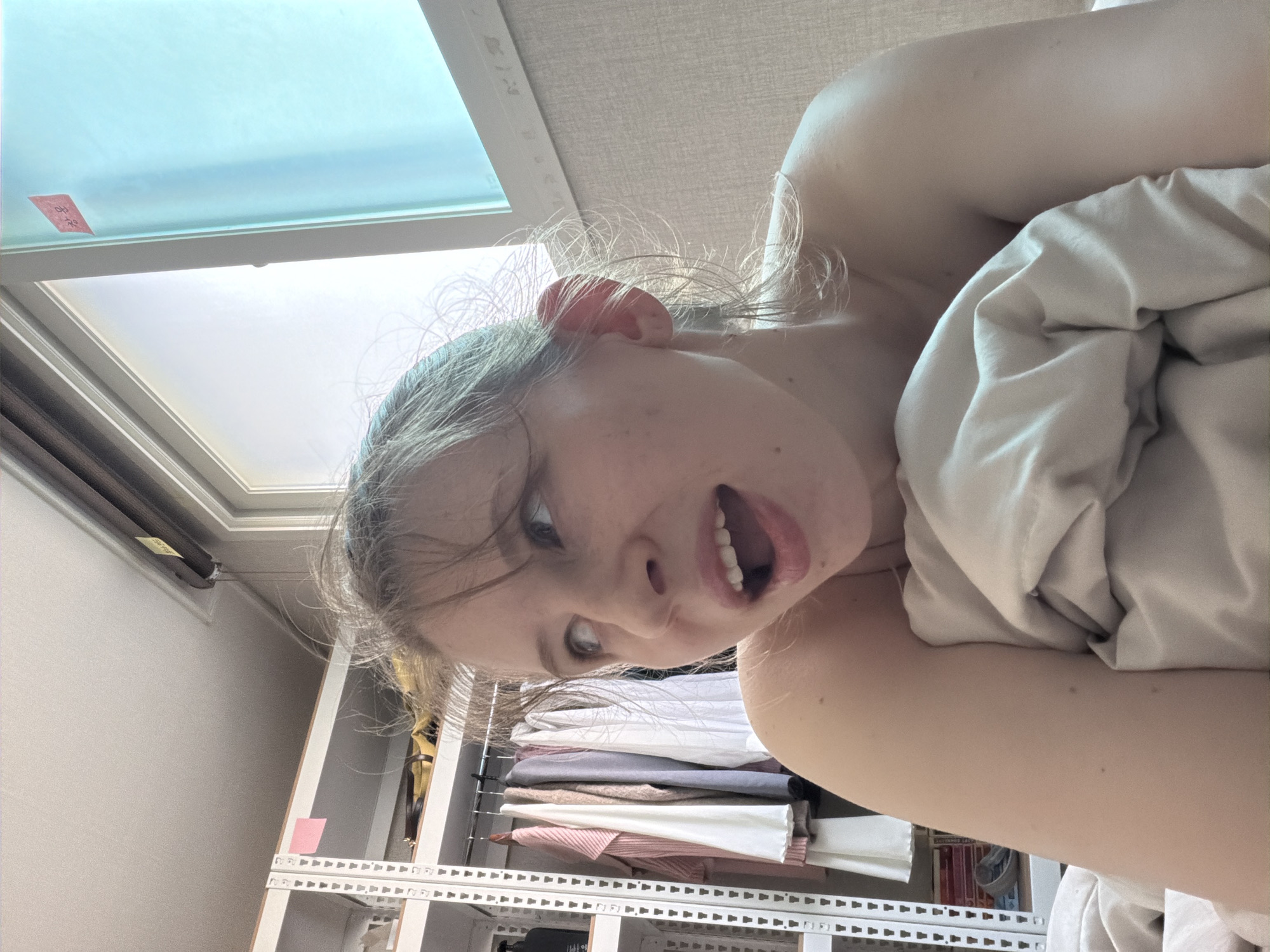 A young Caucasian woman is lying in bed, holding a white comforter around her. She is looking up and to the right with her mouth open, as if surprised or excited. Her hair is messy and light brown. In the background, there is a white clothing rack filled with various garments, a window with a blue tint, and a wall with a textured beige wallpaper. A small red sticker with Korean writing is visible on the window. The overall environment suggests a bedroom in a residential setting, likely within Dongducheon-si, South Korea.