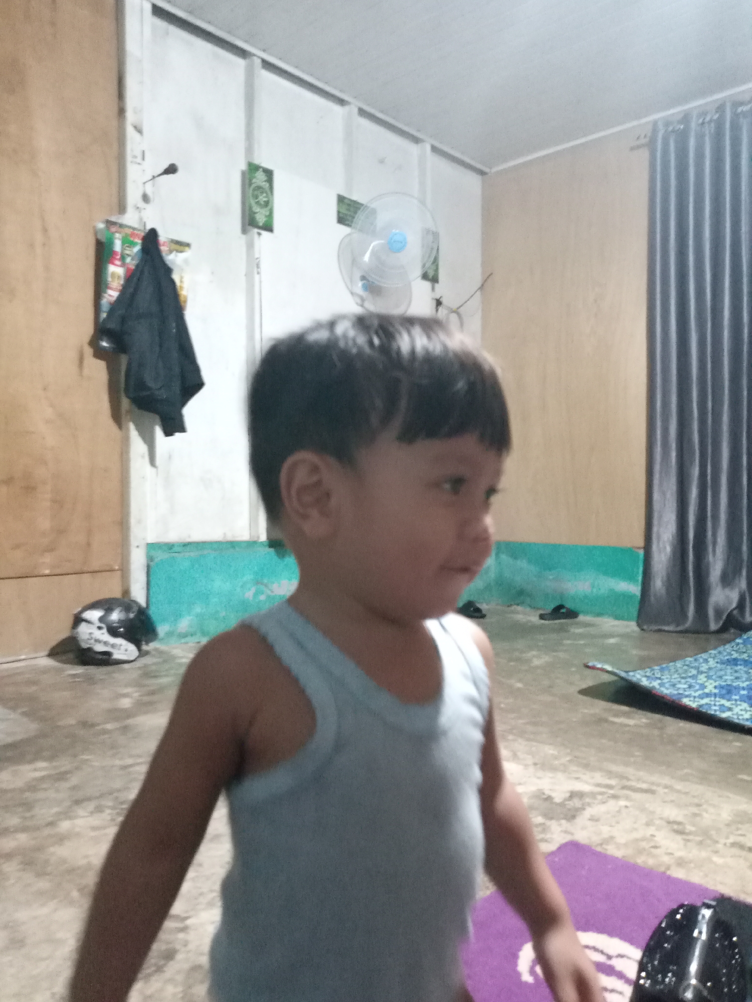 In an indoor setting, a young child in a light blue tank top is captured in a medium shot, facing towards the right of the frame. The child has short, dark hair and a fair complexion. Their head is slightly turned, and their gaze appears to be directed off-camera. The background features a wall with a light-colored upper section and a turquoise lower section. To the left, a wooden door or cabinet is visible, with a black jacket hanging on a hook and some items on a shelf above. A motorcycle helmet with white and black detailing rests on the floor near the wall. Further to the right, a grey curtain hangs, and a ceiling fan is mounted on the wall. The floor is concrete, with a patterned mat in the lower right quadrant of the image. The lighting suggests an indoor environment with artificial light.